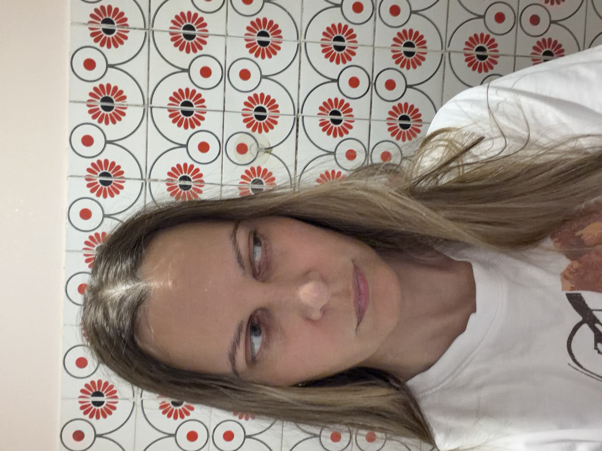 A woman with light brown hair and fair skin is facing the camera. She is wearing a white t-shirt. The background consists of white tiles with a red and black floral pattern. The image was likely taken indoors. There is no evidence to suggest the location is Izhevsk, Russia.