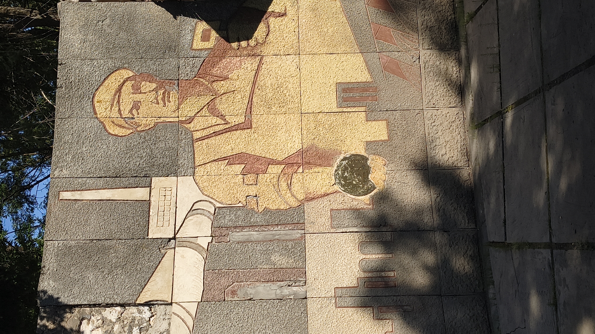 The image shows a mural on a concrete wall, located in Mikhaylovka, Russia. The mural depicts a man in work attire, possibly a factory worker, holding a spherical object in his hand. Behind him, there are industrial elements such as a smokestack and pipes, along with a cityscape. The mural is rendered in a stylized, somewhat abstract manner, with the man's face and clothing textured. The wall itself is constructed of large, rectangular concrete blocks. The ground in front of the wall is paved with large concrete slabs, and shadows from trees are cast across the scene, indicating an outdoor setting. The overall impression is one of socialist realism art, common in Soviet-era public spaces.