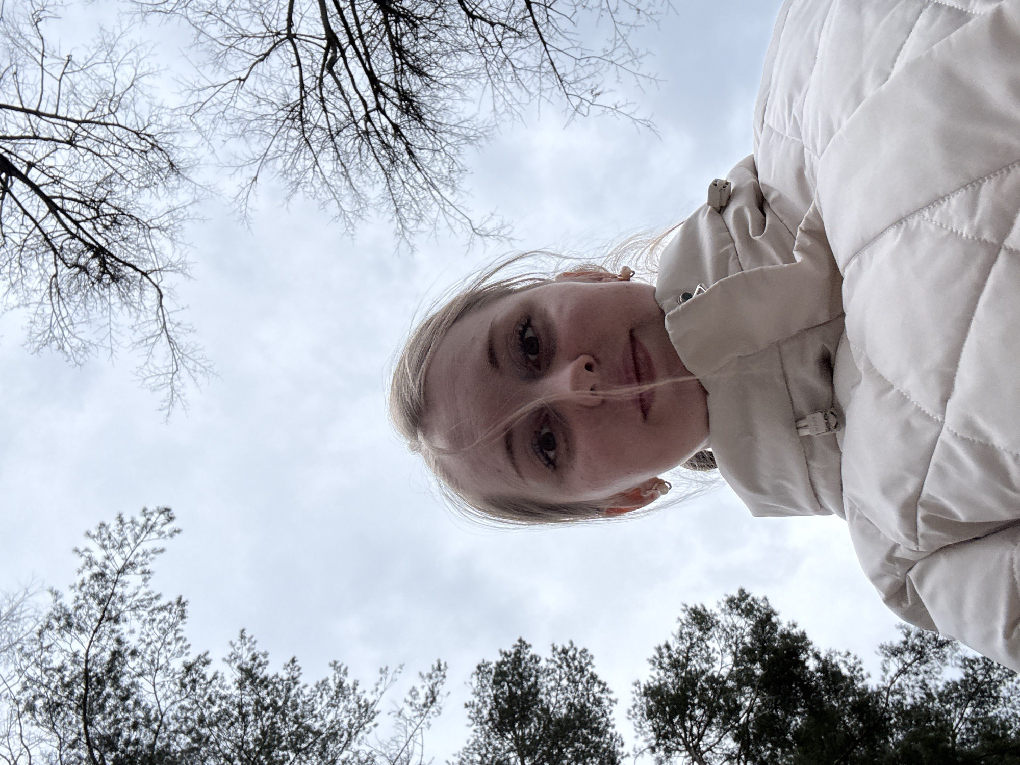 A woman stands outside against a backdrop of a cloudy sky and trees. She wears a light-colored, quilted jacket. Bare tree branches are visible in the upper left of the frame, while green trees are along the bottom.