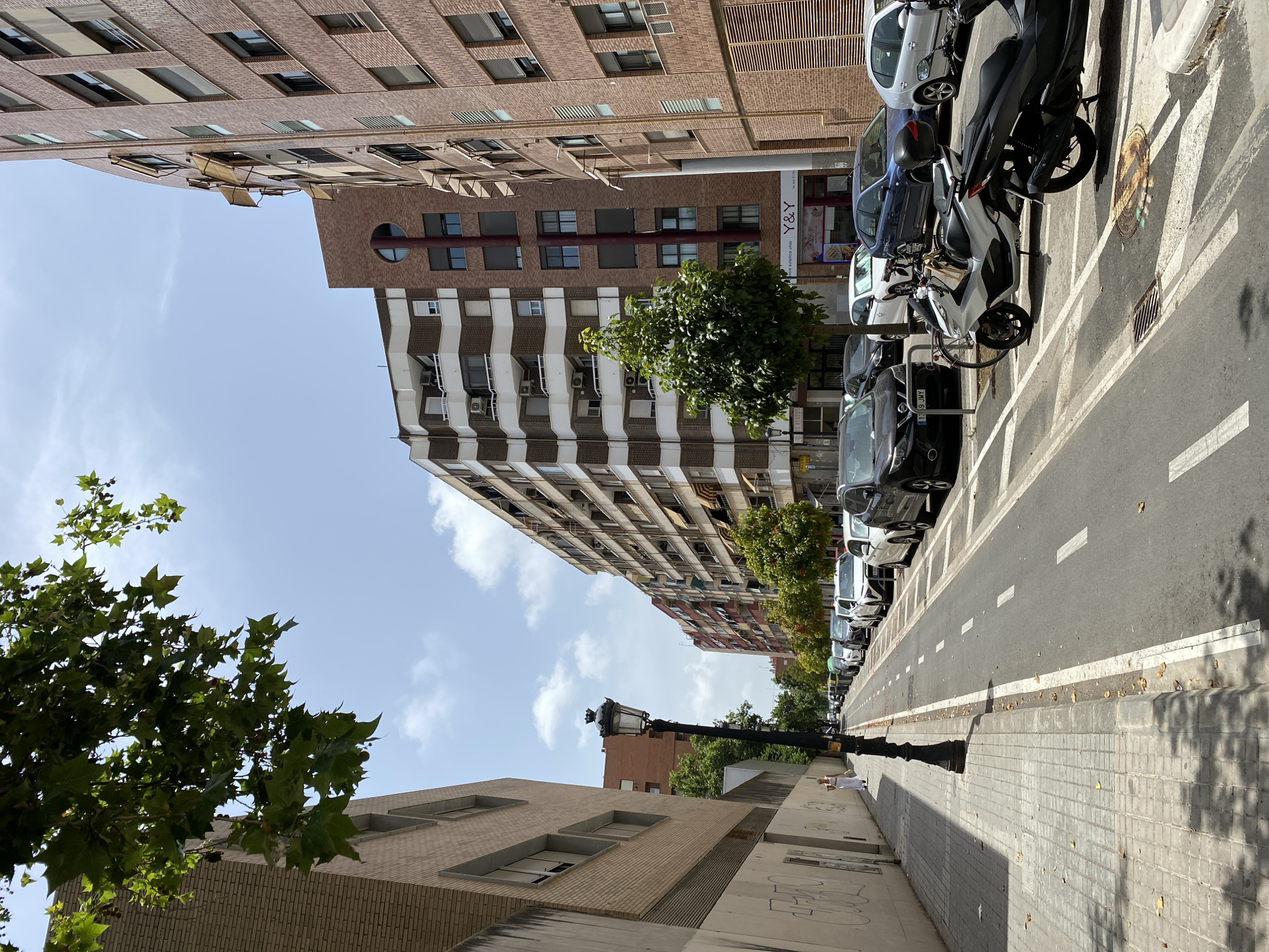 The image depicts a sunlit street in Valencia, Spain, likely on a warm day given the clear blue sky with scattered clouds and the presence of awnings on the buildings. The perspective is looking down a street lined with multi-story apartment buildings constructed with brick and concrete.
On the left side of the street, a paved sidewalk is visible, featuring a light-colored, tiled surface with a section of grey concrete blocks. A single lamppost with a decorative black lantern stands on the sidewalk. A woman in a white outfit is walking on the sidewalk away from the camera. The wall adjacent to the sidewalk has graffiti.
The street itself is a two-way road with a dedicated lane for bicycles marked by white dashed lines. On the right side of the street, parked cars are lined up along the curb. These include various models of sedans and SUVs, as well as two scooters parked in designated spots. The vehicles are parked in an organized manner.
The buildings on either side of the street are tall, with multiple windows and balconies. Some windows have retractable awnings deployed. The building on the right features a rounded facade. A business with a sign reading "Y & Y" is visible on the ground floor of one of the buildings. Trees with green foliage are planted along the street, providing some shade. The overall impression is an urban residential street scene.