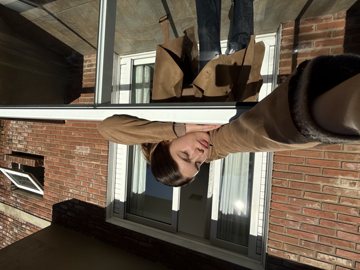 One light-skinned female subject, approximately 18-25 years old, is depicted in a selfie taken on a balcony in Canela, Brazil. She is dressed in a brown, possibly suede, jacket with a knit layer visible underneath, and dark trousers. The subject leans forward, resting her forearms and head on the white top rail of a balcony railing, with her eyes closed or partially shut, appearing to face strong sunlight. Her left arm extends towards the camera. The immediate background features a reddish-brown brick wall. Directly behind the subject is a white-framed double glass door or window unit reflecting intense sunlight. To the right, a white-framed casement window is partially open on an adjacent brick facade. The balcony floor is tiled. Strong, direct sunlight from the left casts distinct shadows across the scene.