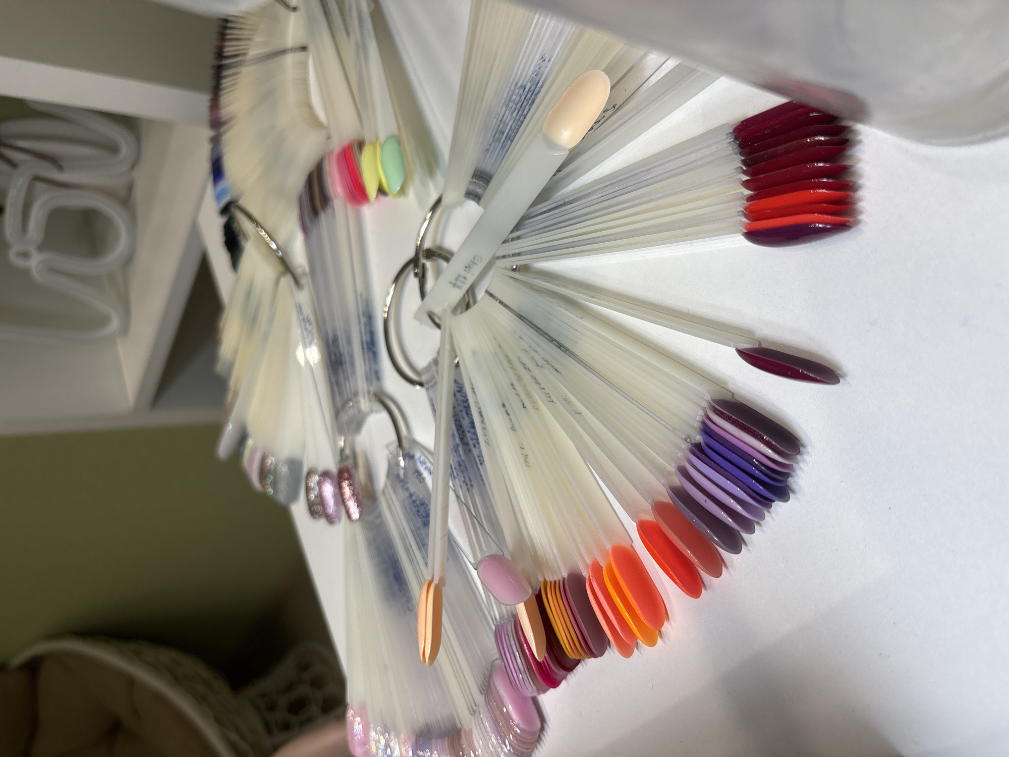 The image shows a collection of nail polish color samples displayed on plastic sticks, arranged in a fan-like manner and held together by metal rings. The colors range from pale nude to bright red and various shades of purple and pink. The samples are in what appears to be a salon or retail setting, given their presentation. The background includes a white shelf with hangers. The location is Ферма, Russia, but no specific contextual details related to that location are visible.