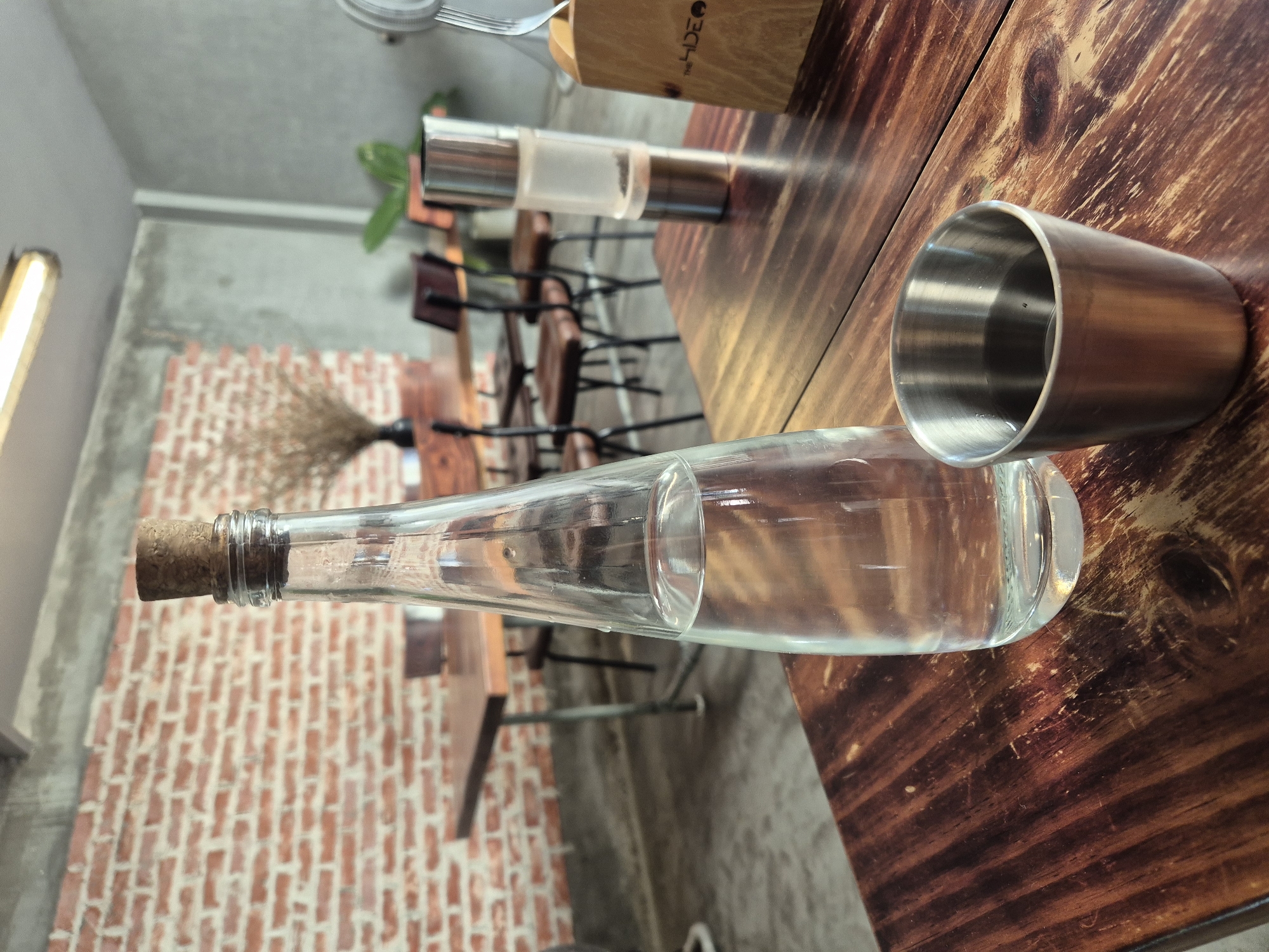 A glass bottle of water with a cork stopper and a small metal cup filled with water are placed on a dark wooden table in what appears to be a cafe. In the background, a brick wall and a concrete wall are visible, along with several wooden tables and chairs. A pepper grinder and a wooden menu holder with the text "THE SIDE" are also on the table. The overall ambiance suggests a casual dining or coffee shop setting. The location is a cafe in Đà Nẵng, Vietnam.