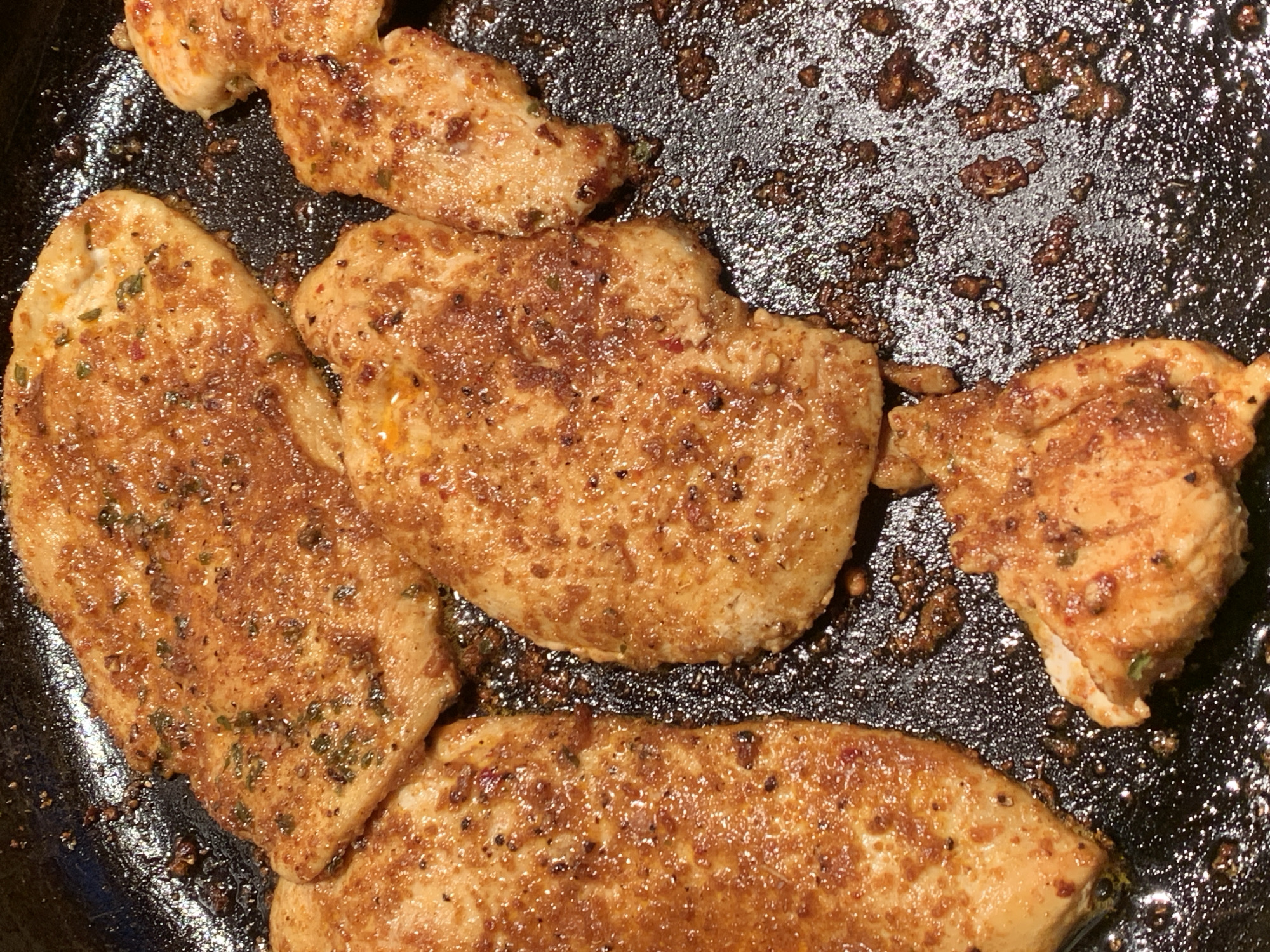 In a close-up, overhead shot, several pieces of seasoned chicken breast are seen sizzling in a black, non-stick frying pan. The chicken pieces are thinly sliced and coated in a reddish-brown spice mix that includes visible flecks of herbs and red pepper flakes. The pan is dark and appears to have accumulated some rendered fat and browned bits from the cooking process, creating a glossy, slightly textured surface around the chicken. The lighting is warm, highlighting the golden-brown sear on the chicken. There is no indication of the location being in Barcelona, Spain; it appears to be a domestic kitchen setting.