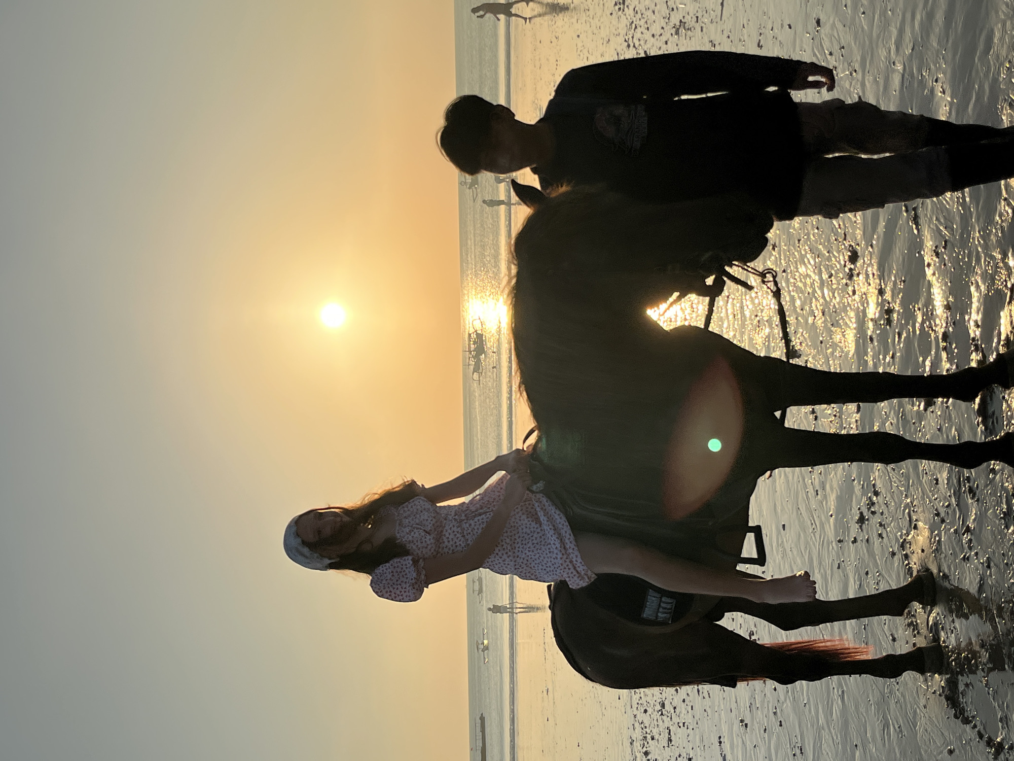 At sunset on Kuta Selatan beach, Indonesia, a young woman with a white headscarf rides a dark brown horse on the wet sand. She wears a short-sleeved white dress with a floral pattern. To her right, a young man stands by the horse's head, wearing a dark long-sleeved shirt and light-colored cargo pants. The sun is low on the horizon, casting a warm golden glow across the scene and reflecting on the water and wet sand. Several small boats are visible in the calm ocean in the background, and other people can be seen walking on the beach further in the distance. The tide is low, revealing a rippled sand surface. A lens flare, appearing as a green circular light, is present on the horse.
