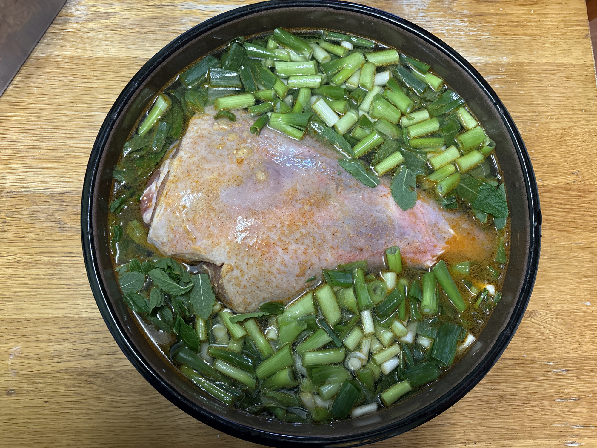 A top-down view shows a dark, circular bowl filled with a broth-like liquid, containing a large cut of raw meat, possibly pork or poultry. The meat is pale in color with some reddish-orange seasoning on its surface. Chopped green onions and mint leaves are scattered throughout the broth. The bowl sits on a light brown wooden surface. There is no evidence indicating the location is Oswestry, United Kingdom.