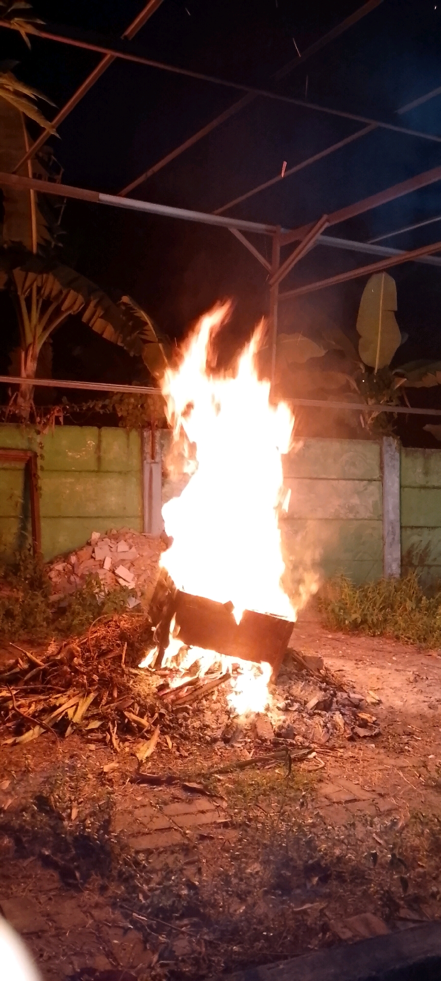 A nighttime outdoor scene in Satriamekar, Indonesia features a bonfire. The fire is contained within a dark, rectangular metal structure, possibly a drum or bin, which is tilted and sits on a pile of debris that includes dry leaves and branches. The flames are bright white and yellow, reaching high into the air, illuminating the surrounding area with an orange glow. Behind the bonfire, a light green wall made of concrete blocks stretches horizontally. To the left of the wall, a large banana tree with broad leaves is visible. Above the bonfire and wall, a metallic frame structure, possibly part of a roof or shelter, is silhouetted against the dark night sky. The ground in the foreground appears to be a mix of dirt, gravel, and scattered debris. There are no people visible in the image.