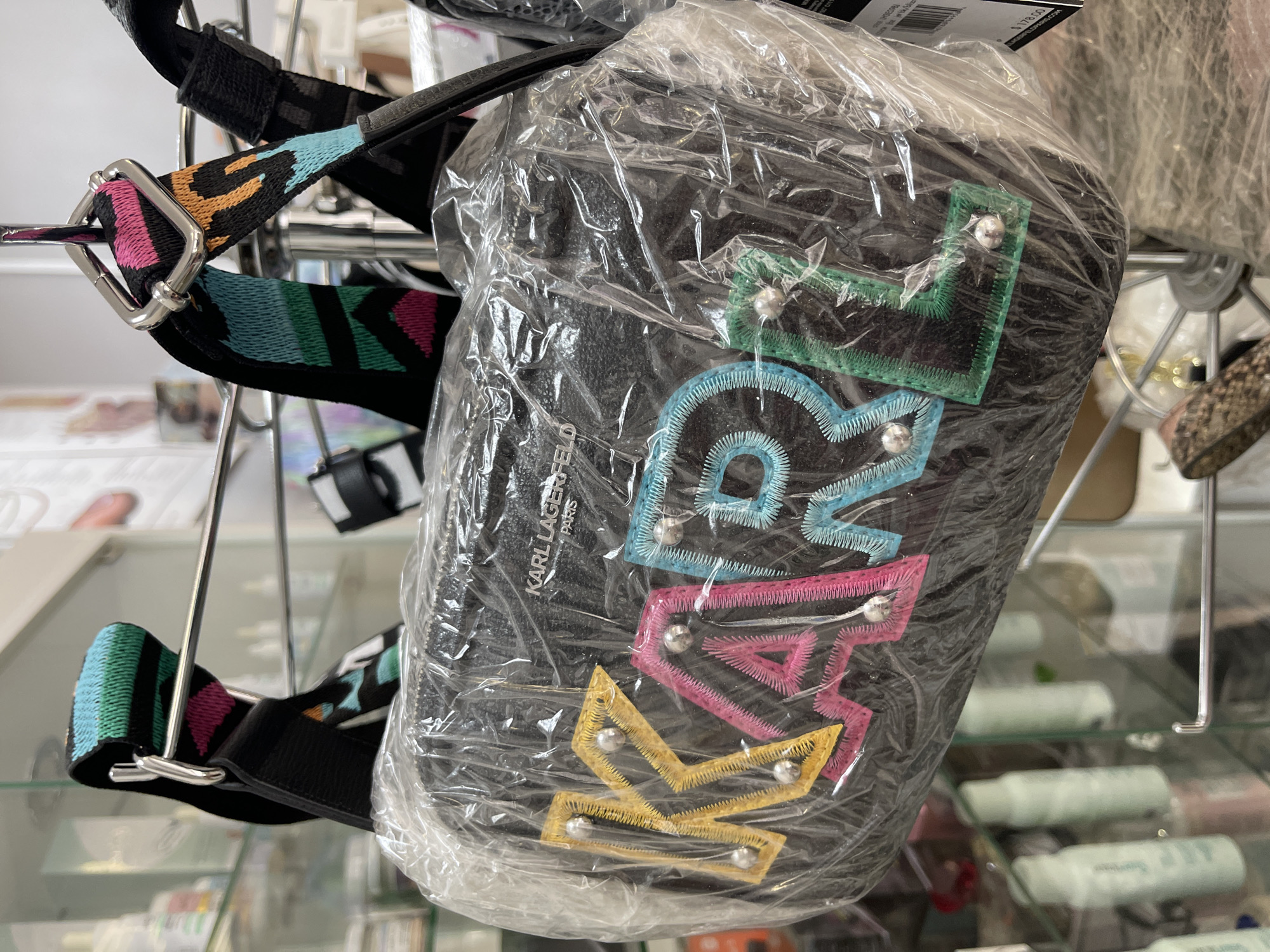 A black Karl Lagerfeld Paris bag, covered in clear plastic, hangs on a metal rack. The bag has a colorful, embroidered design of four overlapping crowns stacked vertically: the top crown is green, followed by blue, pink, and yellow. Each crown has a small, silver-colored stud at its points. The bag is hanging by a colorful strap with a black leather attachment. The background shows a retail environment with shelves and other items. The scene is lit by overhead artificial light. The image does not reveal the location or city.