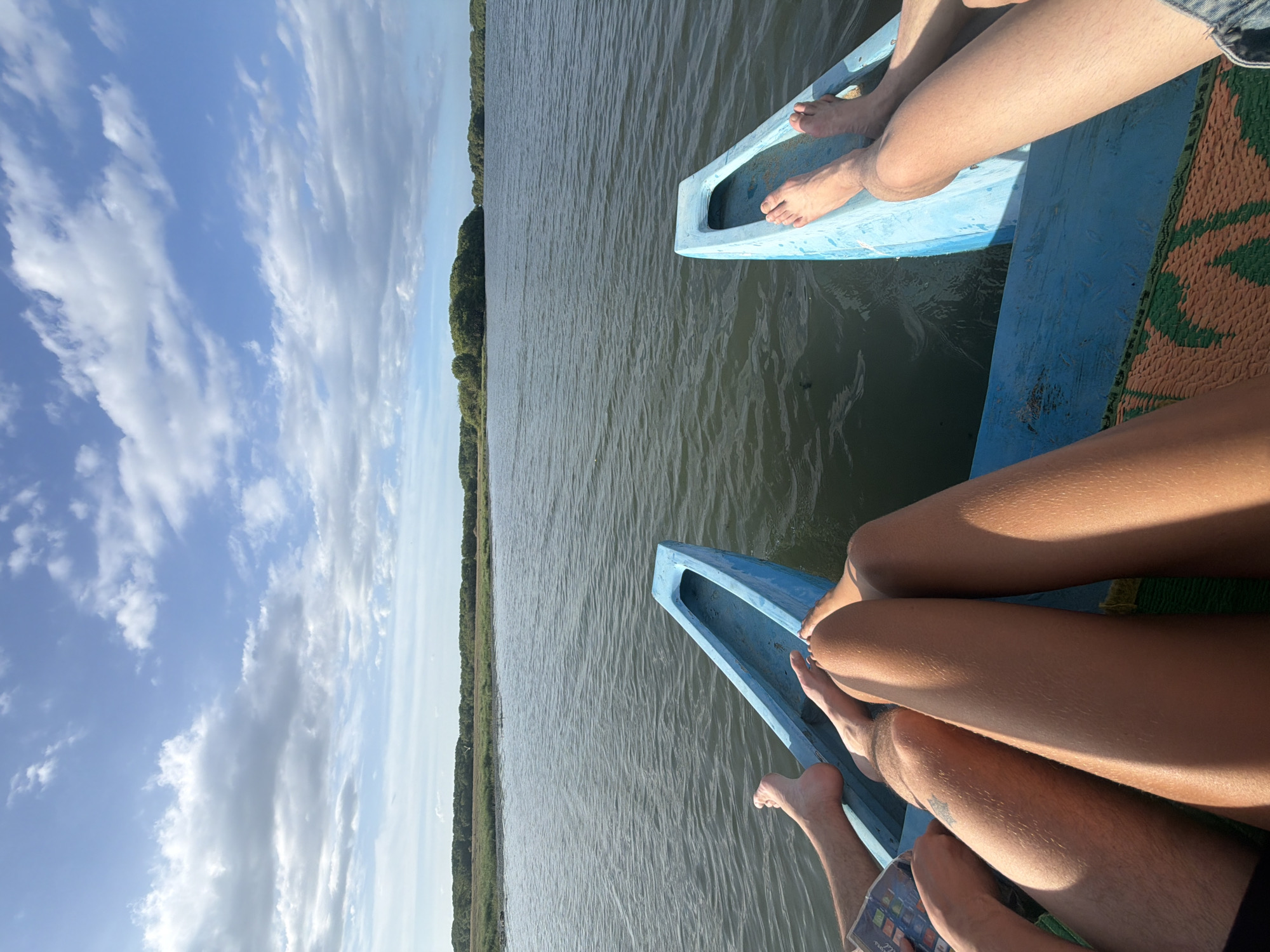 This image captures a serene moment from a boat journey on a body of water, likely a lagoon or river, in Pottuvil, Sri Lanka. The perspective is from within the boat, looking forward. Two light blue outriggers of a traditional boat are visible, one on the left and one on the right, extending towards the viewer. The water is calm with subtle ripples, reflecting the overcast sky.
In the foreground, the legs and feet of at least three individuals are visible, resting casually within the boat. One person's legs are crossed, another pair of legs are extended forward. The boat appears to be adorned with a woven mat in shades of orange and green.
The background features a lush, green shoreline densely populated with trees, indicative of a coastal or riverine environment. The sky above is a mix of blue and scattered white clouds, suggesting it is daytime, possibly late afternoon given the lighting. The overall impression is one of relaxation and immersion in nature, typical of a tranquil excursion in the scenic region of Pottuvil.