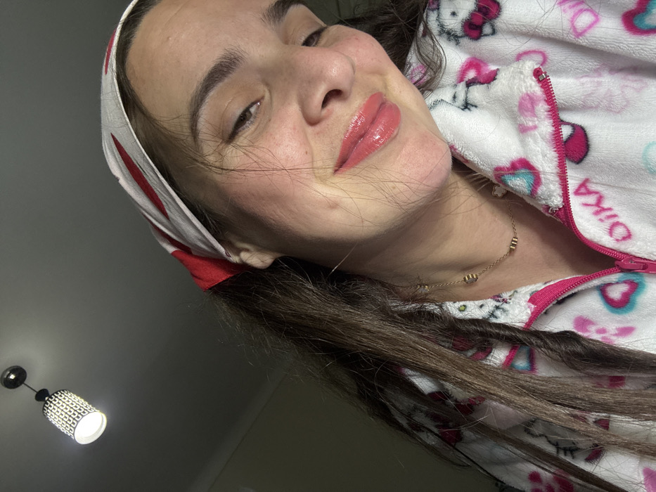 A woman tilts her head towards the camera, sporting glossy lips and a playful smirk. She wears a white headband with red accents and a zippered pajama top patterned with pink and blue designs over what appears to be Hello Kitty imagery. A delicate gold necklace adorns her neck. Dark hair frames her face and cascades downward. A modern hanging lamp provides illumination from the bottom left corner of the image.