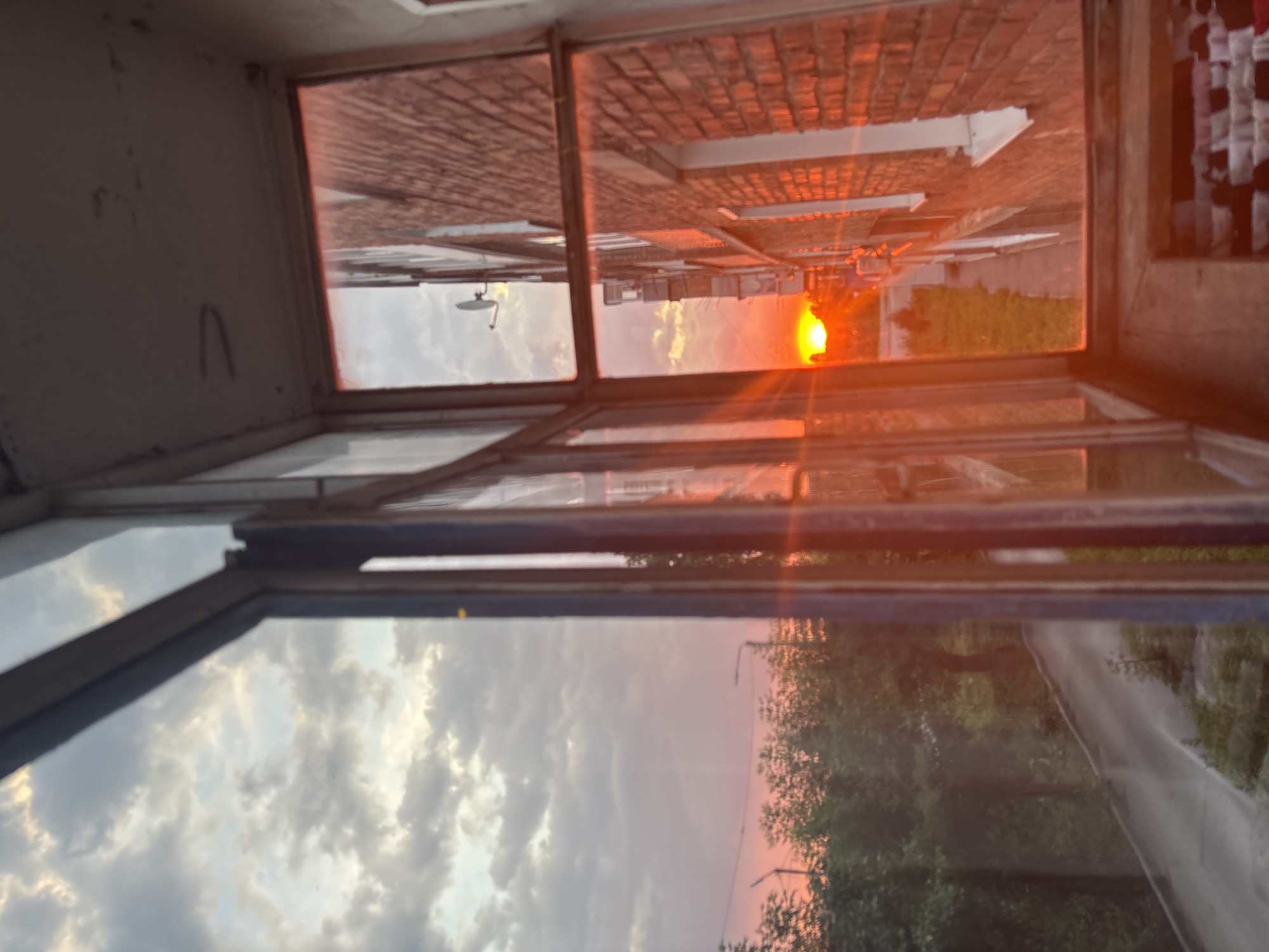 The image depicts a view through a window frame looking out at a sunset. The framing of the image suggests an indoor perspective, possibly from a balcony or a room with a window. The scene outside is dominated by a vibrant sunset with the sun appearing as a bright, orange orb. The sky is filled with dramatic clouds, illuminated by the setting sun, creating a warm, fiery glow.
The exterior view appears to be a street or pathway bordered by buildings on one side and trees on the other. The buildings have brick facades, and there are visible structural elements like window frames and possibly rooflines. The trees show green foliage, suggesting a temperate climate. The presence of streetlights indicates an urban or suburban setting.
The image is characterized by strong reflections on the glass, adding a layer of complexity to the scene. These reflections include parts of the interior environment, such as the wall and ceiling, as well as more of the exterior, further blurring the lines between inside and outside. The overall impression is one of a peaceful, possibly contemplative moment, captured during the transition from day to night. The warm colors of the sunset evoke feelings of warmth and tranquility. The image's orientation is rotated, with the sunset appearing at the top and the sky at the bottom, suggesting the original capture was not upright. No visible text is present in the image.
