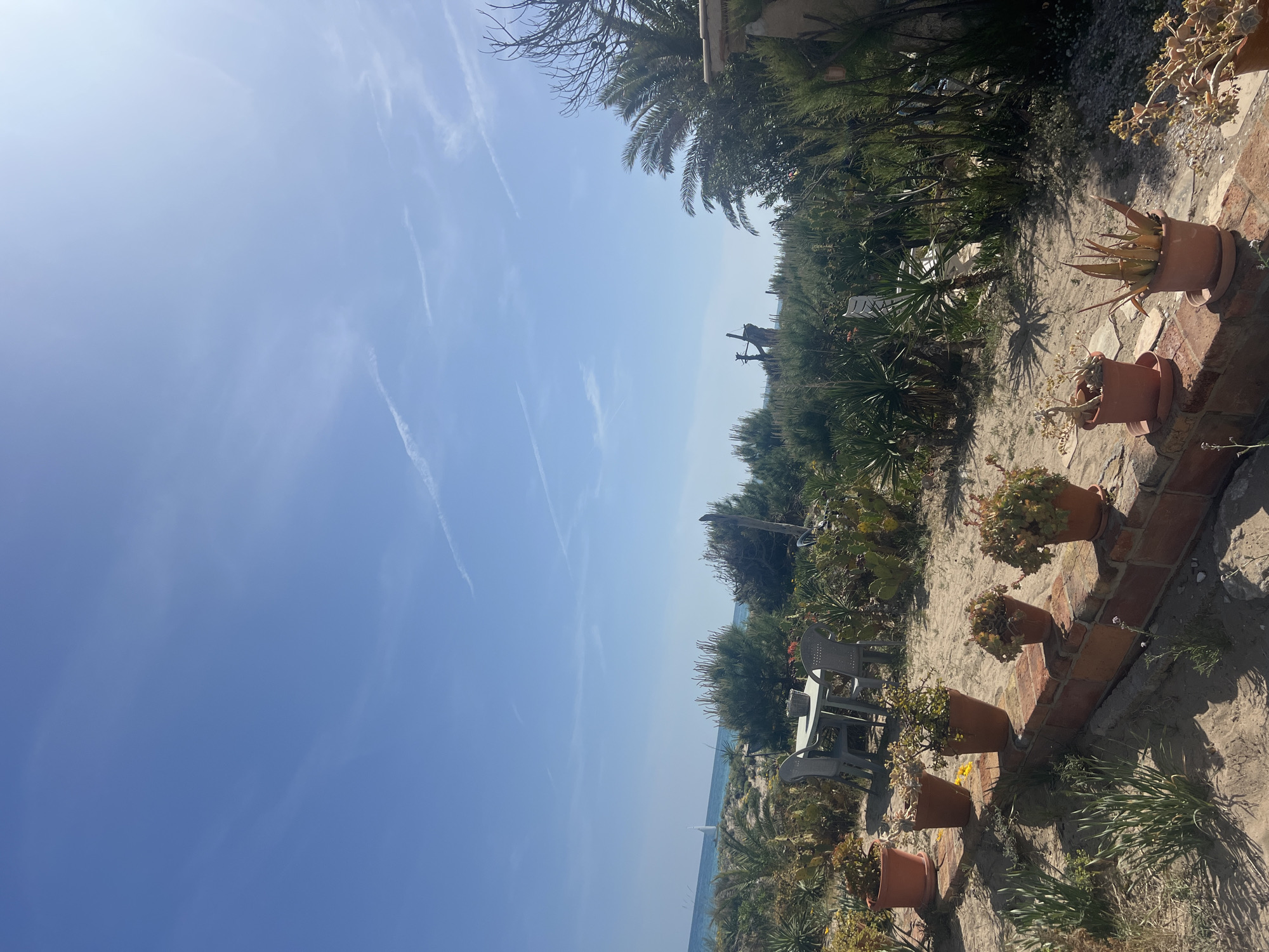 The image captures a coastal scene in La Marina De Creixell, Spain. A clear blue sky with faint contrails provides the backdrop. Vegetation, including palm trees and various shrubs, lines the edge of a sandy path. Terracotta pots containing plants are arranged along a brick wall or structure. In the distance, a white table and chairs are visible near the coastline. A sailboat is discernible on the water.