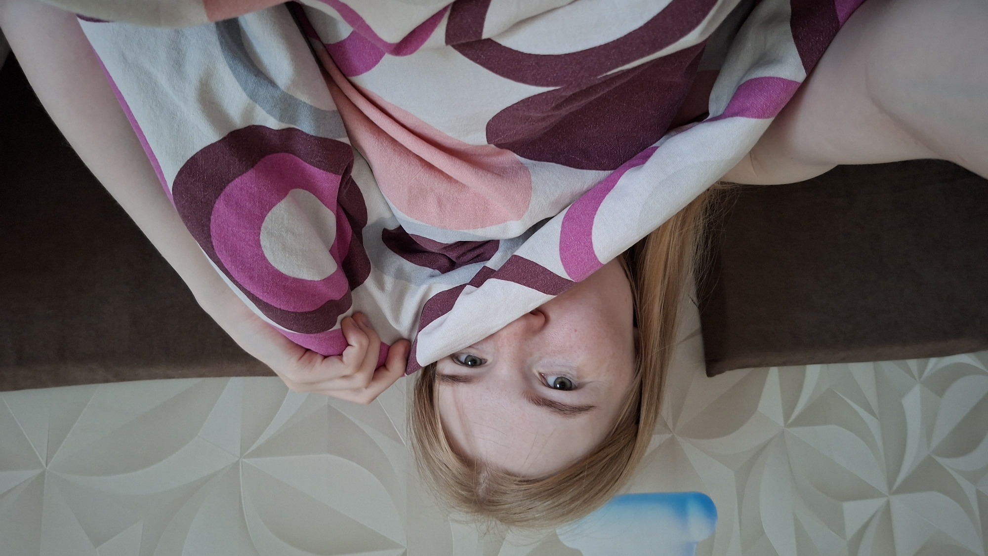 In a bedroom, a person with blonde hair is partially covered by a duvet with a circular pattern in shades of pink, maroon, and white. The person's face is visible from the eyes down, and one hand is holding the duvet near their face. The background includes a textured wall with a cloud decal and a dark brown headboard. The image does not provide specific details to determine the location within the city of Chaikovsky, Russia.