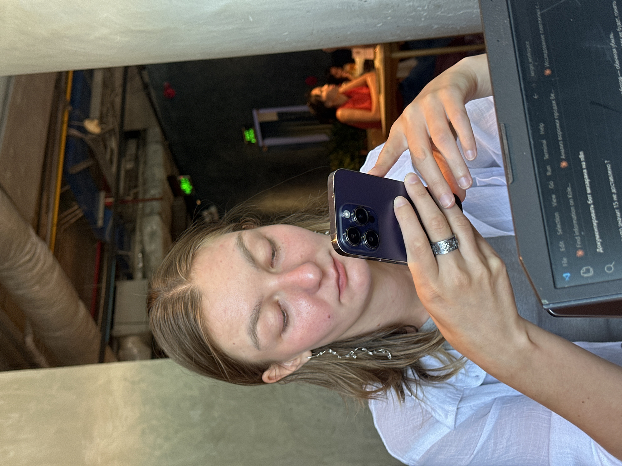 A young woman with fair skin and light brown hair pulled back from her face is seated at a table, looking down at her dark purple smartphone. She is wearing a white collared shirt over a grey t-shirt and silver earrings that hang down past her jawline. A silver ring with an intricate design is on her right hand. Her left hand is also visible, holding the phone. In the foreground, part of a laptop screen is visible, showing lines of code and text in Russian. The background is dimly lit, with another woman in a red shirt visible at a table in the distance. The overall setting appears to be an indoor cafe or restaurant. The specific location in Da Nang, Vietnam, cannot be determined from the image.