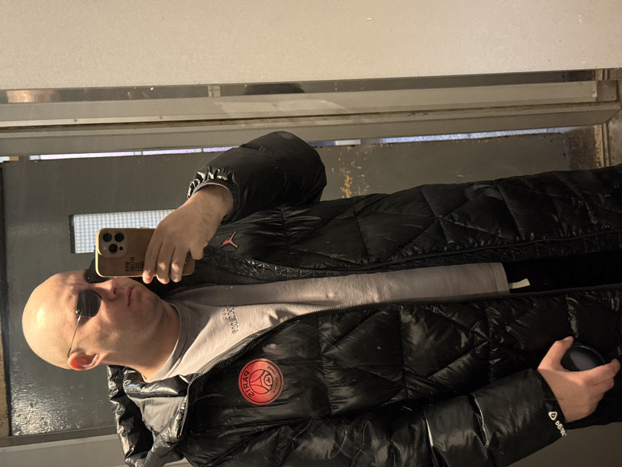 An adult male with a bald head and dark, frameless rectangular sunglasses is depicted taking a mirror selfie indoors. He wears a shiny black quilted puffer coat, open to reveal a grey crew-neck t-shirt underneath. The coat features a red and black circular "PARIS SAINT-GERMAIN" (reversed) patch on the left chest and a small red "Jumpman" logo on the right chest. White "66BK" (reversed) text is visible on the left cuff of the jacket. He holds a light brown smartphone with three prominent camera lenses in his raised right hand, pointed towards himself. A small, dark, circular object is held in his left hand. The immediate background, reflected in the mirror, consists of a plain light-colored wall to the viewer's left and a darker panel or door with a translucent or textured glass insert to the viewer's right. A vertical door frame or jamb separates these two background elements. The specific location within the city of Pleven, Bulgaria, is not discernible from the visual evidence provided.