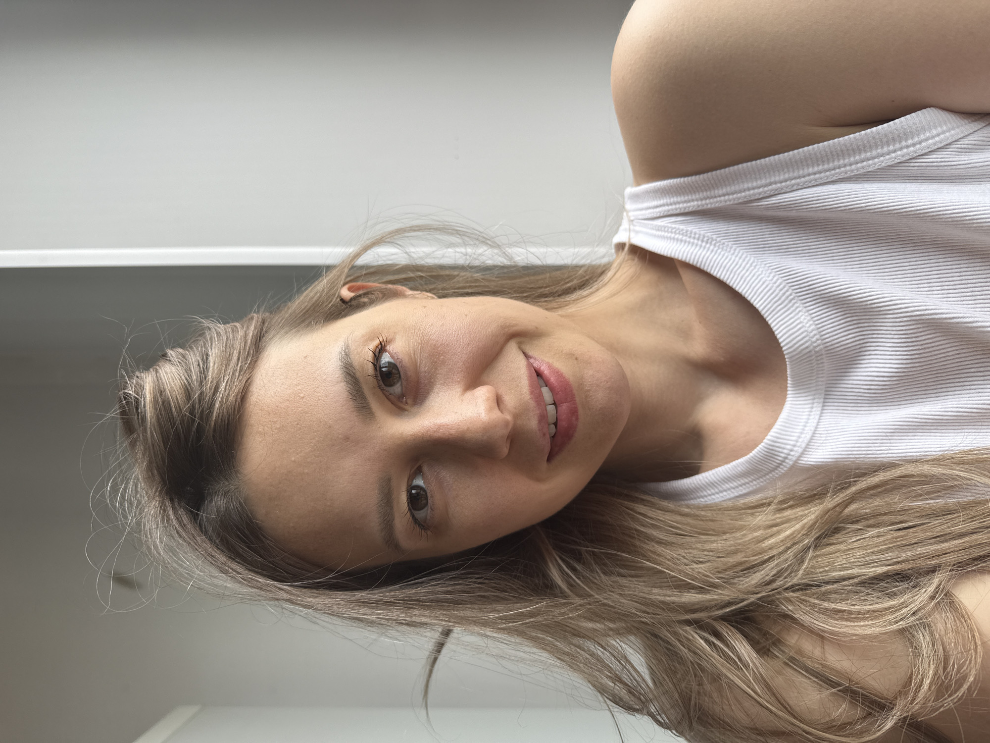 A woman with fair skin, light brown hair, and brown eyes is posing indoors. She is wearing a white ribbed tank top. The background appears to be a white wall, possibly with shelves or cabinets. There is no information to infer the location is in the city of Chaikovsky, Russia.