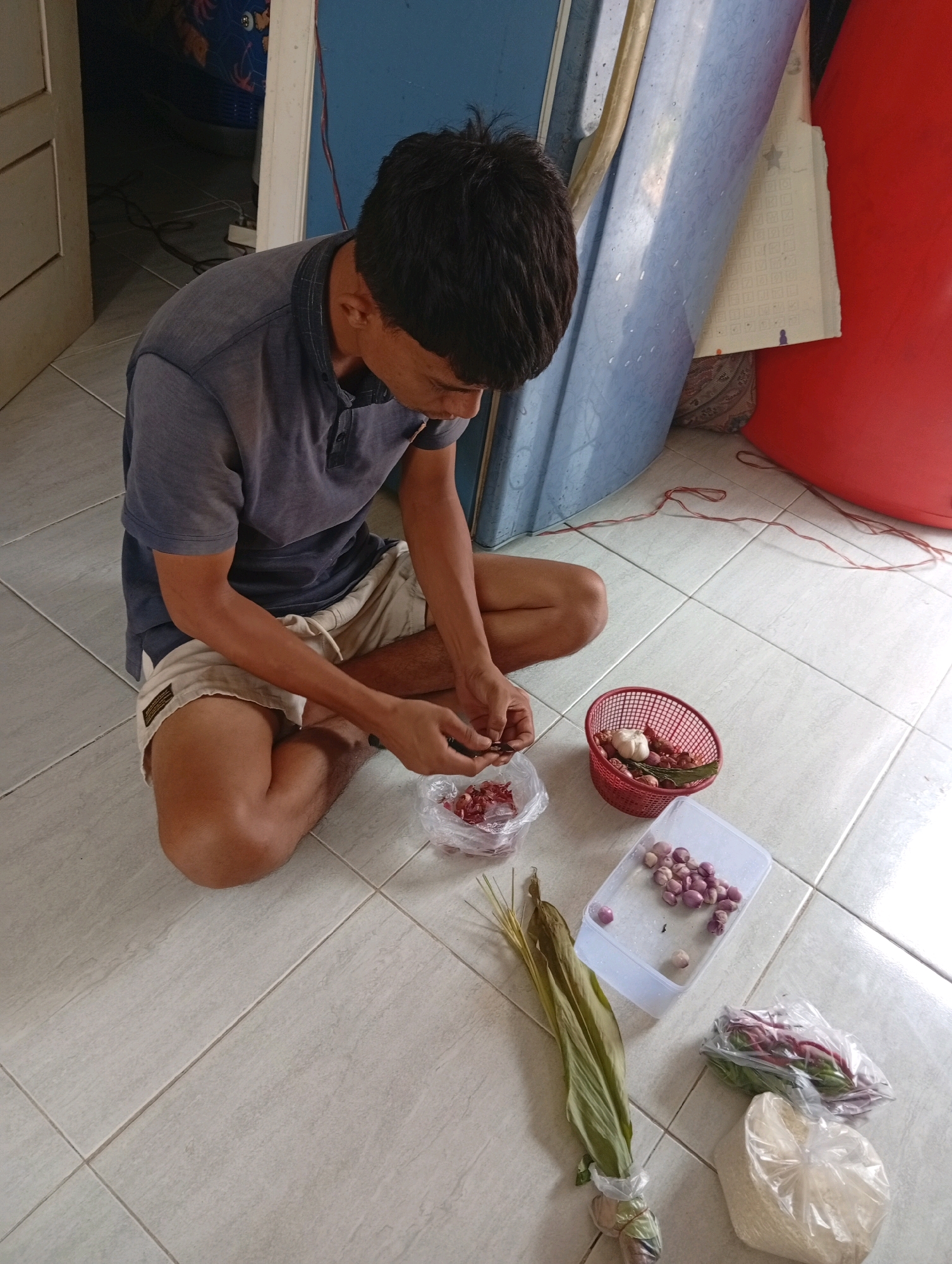In Kualu, Indonesia, a person is sitting on a tiled floor, preparing ingredients. The individual, with short, dark hair and wearing a blue t-shirt and beige shorts, is crouched with their legs crossed. They are actively engaged in peeling or chopping small, reddish items from a clear plastic bag. In front of them, several other food items are laid out: a red basket containing garlic and what appears to be shallots or small onions, a clear plastic container filled with more shallots, a bunch of long, green leafy herbs, a plastic bag with red-stemmed vegetables, and another plastic bag containing a white granular substance, likely rice. The scene takes place indoors, indicated by the tiled floor and the presence of a blue, curved object and a red object in the background, possibly part of furniture or stored items. The lighting suggests an indoor environment, possibly daytime given the natural light.