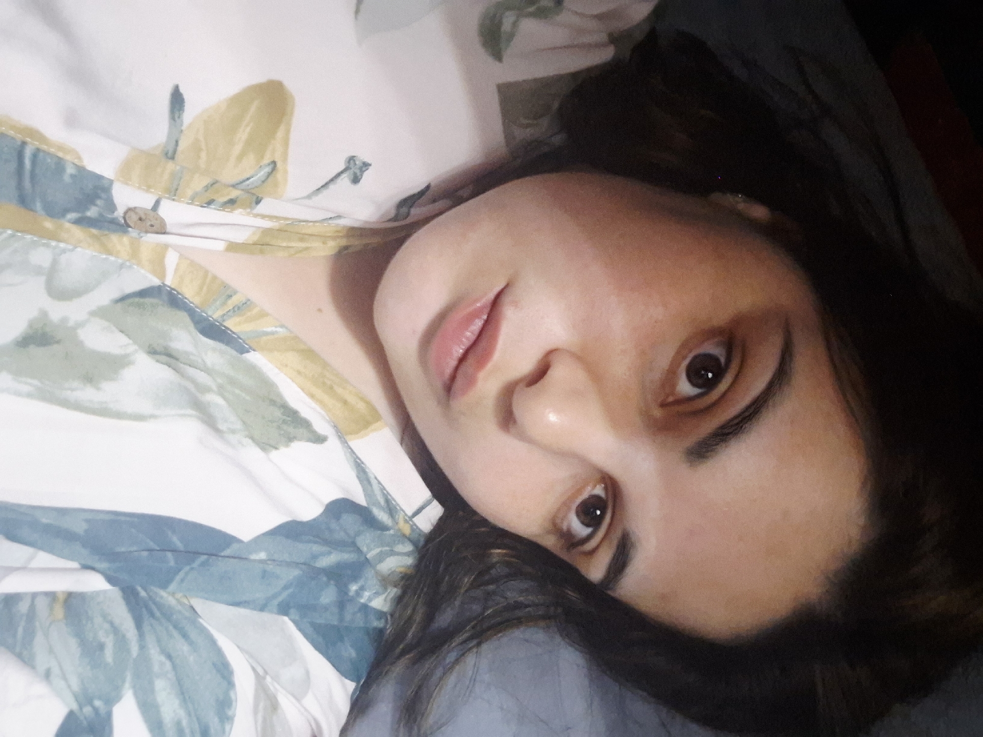 The image shows a woman lying down. She is fair-skinned with dark hair and brown eyes. She is wearing a light-colored shirt with a floral pattern and a button. Her head is resting on a grey surface, possibly a pillow or blanket. The background is blurry and dark, suggesting an indoor setting. It is not possible to determine the location as Амзя, Russia from this image.