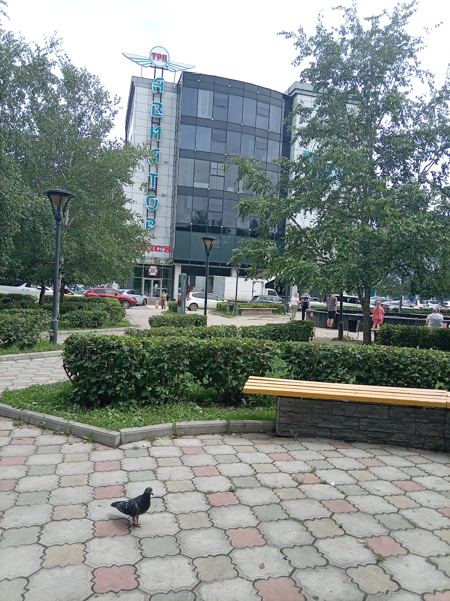 The image depicts the "TseRTS" shopping center in Artyom, Russia. The building features a modern glass facade and a prominent rooftop sign with wings and the letters "ТПЦ". Vertical blue lettering spelling "АВИНАТОР" is also visible on the building's exterior. In the foreground, a paved area with a decorative tile pattern is occupied by a pigeon. Beyond the paving, a well-maintained garden with manicured hedges surrounds a yellow wooden bench. Several people are seen walking near the entrance of the shopping center, and cars are parked in the background. The scene is set on an overcast day, with trees providing shade.