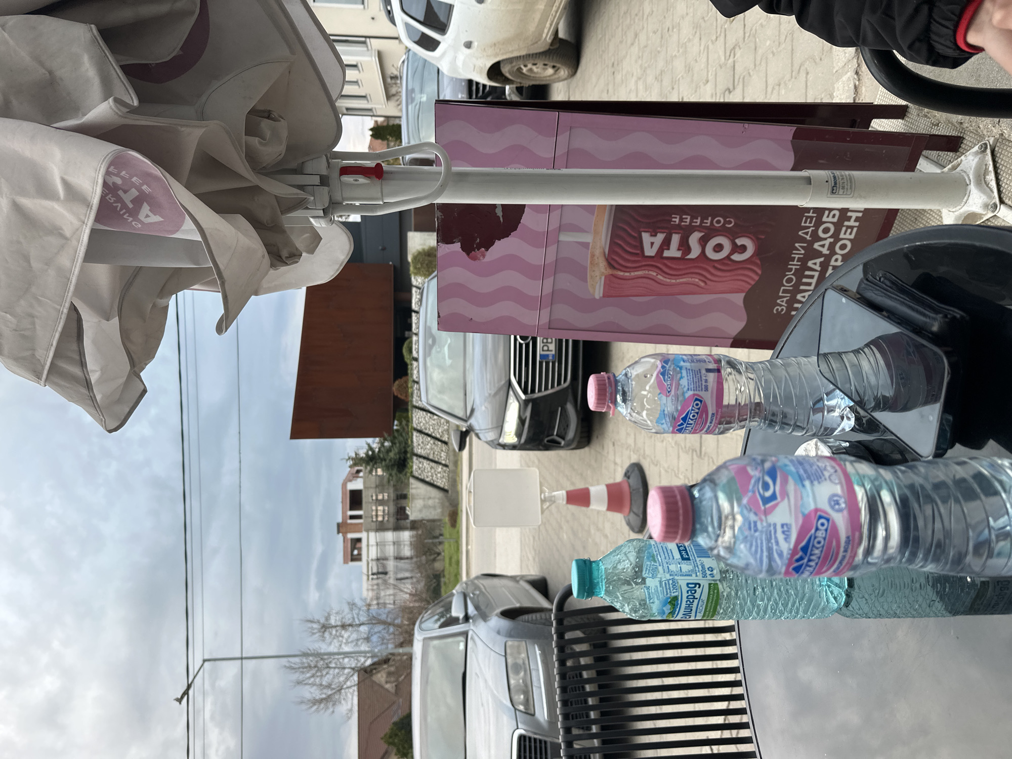 The scene is an outdoor patio setting in Plovdiv, Bulgaria, featuring a Costa Coffee advertisement stand. Three water bottles sit on a glass table with a black metal base. The bottles are labeled "Halkovo," a Bulgarian mineral water brand, and "Velingrad." An open sun umbrella is visible overhead, and behind the scene is a row of parked cars, including a silver Audi, along the street. A traffic cone sits near the bottles. The setting appears to be adjacent to a building complex with scaffolding, under a cloudy sky. A person wearing a black jacket with a red trim is partially visible at the top right corner.