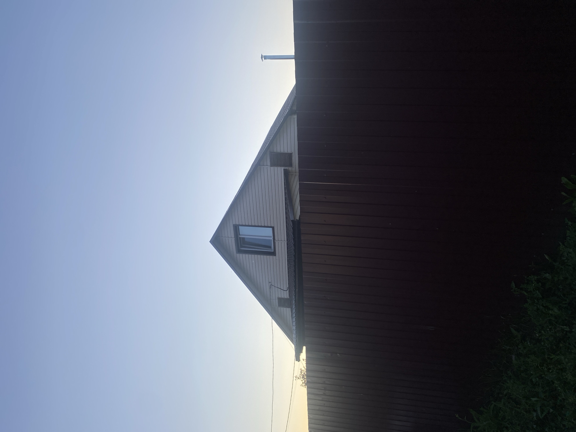 The image depicts a residential scene in Verkhny Karbush, Russia, under a clear, pale blue sky. A two-story house with beige siding and a dark roof is visible. The house has a prominent gable roof with a single window on the upper floor, which appears to be open. A metal chimney extends from the roof. A dark brown, corrugated metal fence runs across the foreground, partially obscuring the lower portion of the house. Green vegetation is visible at the bottom left of the image, in front of the fence. The overall lighting suggests either early morning or late evening.