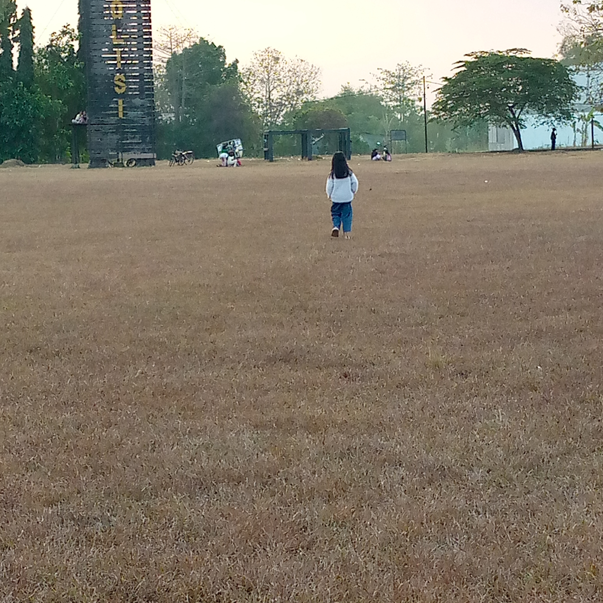 The image depicts a wide, dry, grassy field under a bright sky, likely during the day. In the foreground, a young child, seen from behind, walks away from the camera. The child wears a light-colored long-sleeved shirt and blue pants. In the background, a tall, dark, wooden structure with yellow markings is visible, possibly a tower or monument. To the left of this structure, some trees and foliage are present. Further in the background, to the right of the child, a large, leafy tree stands prominently. Other indistinct figures and objects are scattered across the field, including what appear to be bicycles and people sitting or gathered in small groups. The overall environment suggests an outdoor recreational area or park. The location is identified as the Pacing city in Indonesia.