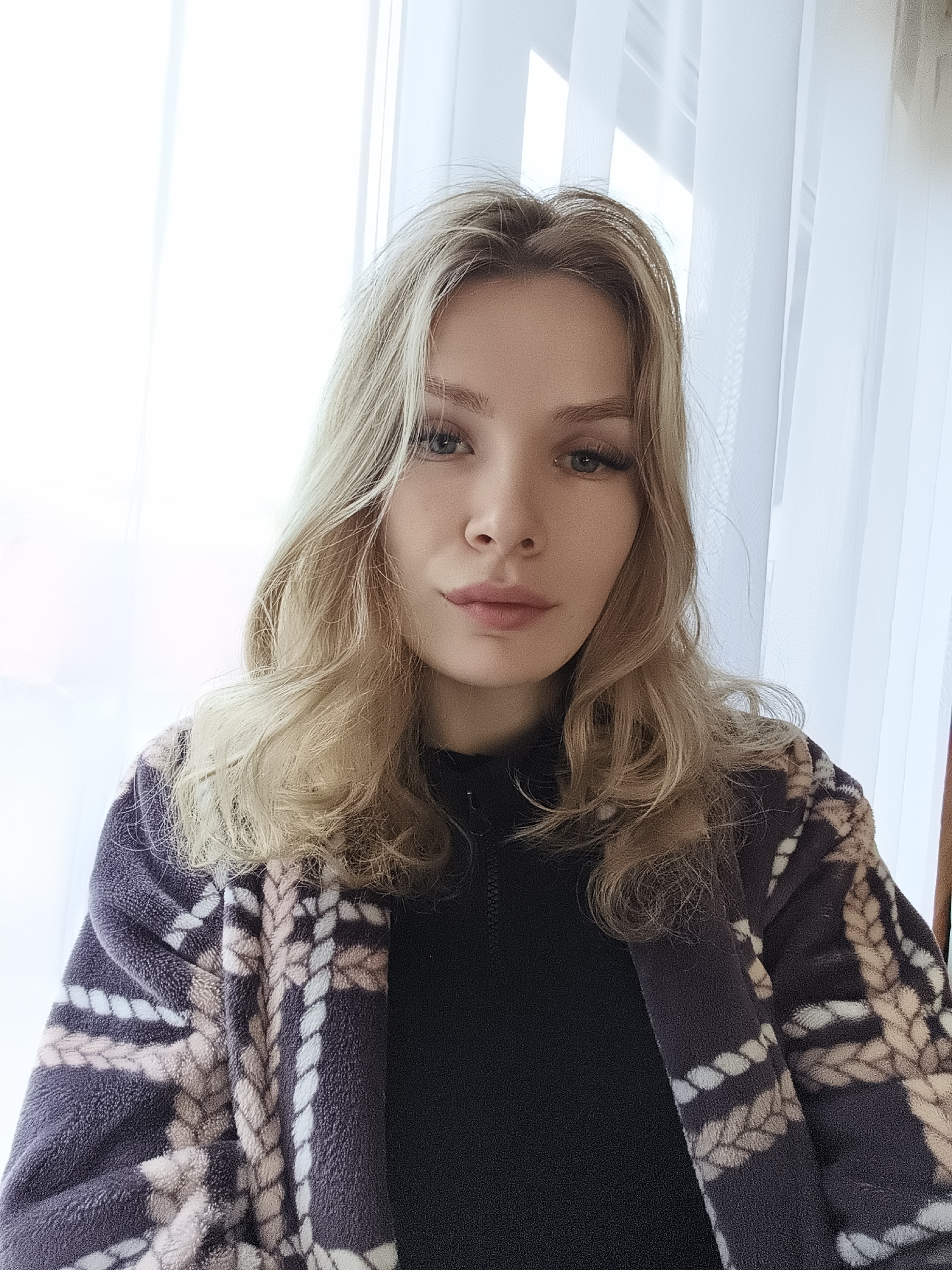 A fair-skinned woman with long blonde hair is looking directly at the camera. She has light eyes and is wearing false eyelashes. Her lips are slightly parted. She is wearing a black top and a patterned robe or jacket in shades of grey, beige, and white. The pattern appears to be a rope-like design. Behind her, there are sheer white curtains covering a window. The background is brightly lit. There is no identifiable information to confirm the location of Русский Вожой, Russia in this image.