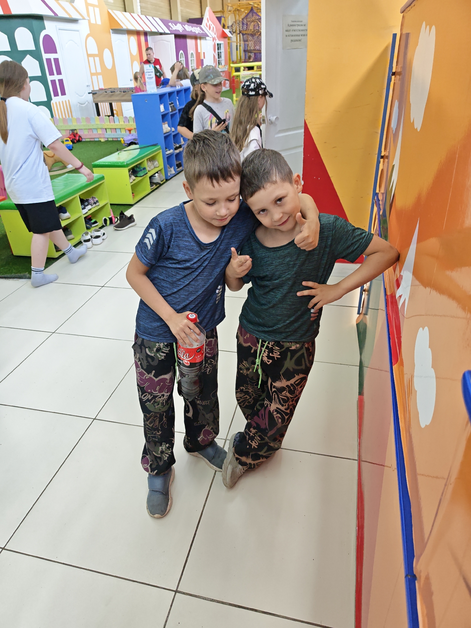Two young boys are posing for a photo indoors. The boy on the left is wearing a blue shirt and camouflage pants, holding a large bottle of Coca-Cola. He is leaning slightly towards the boy on the right, who has his arm around him, giving a thumbs up. The boy on the right is wearing a green shirt and similarly patterned camouflage pants. He is leaning against a brightly colored orange and blue wall. The background shows an indoor playground with miniature buildings and children's activities. There are other children in the background near a shoe rack. The location is Chaikovsky, Russia.
