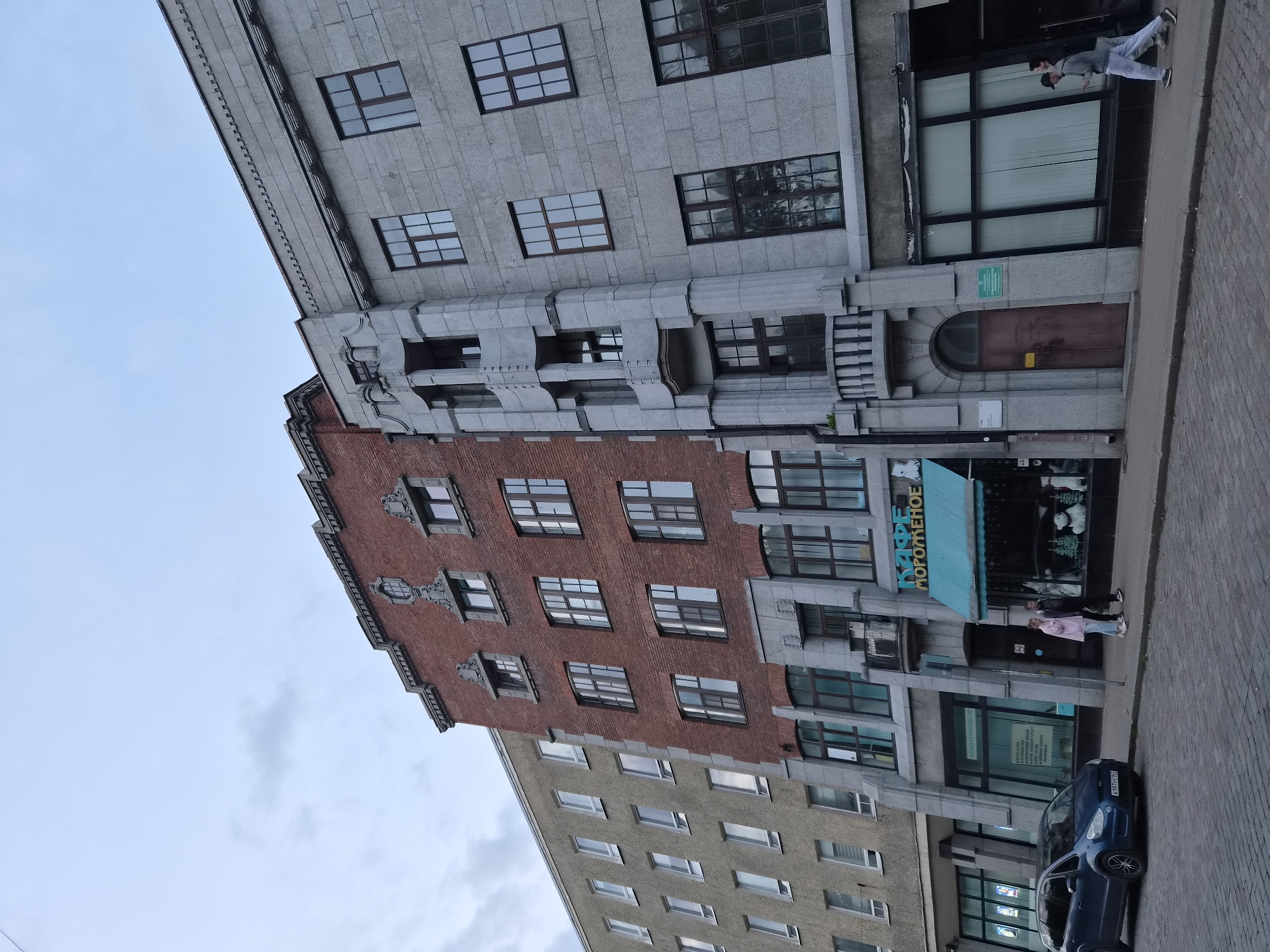 The image shows a street scene in Vyborg, Russia, featuring a row of multi-story buildings with varying architectural styles. The leftmost building is light gray and has a repetitive pattern of windows. Adjacent to it is a brick building with darker window frames and decorative stone elements around some of the windows. The rightmost building is constructed of gray stone blocks and features a prominent stone facade with a curved, recessed section containing windows and a balcony.
At street level, there is a storefront with a teal awning and a sign that reads "КАФЕ МОРОЖЕНОЕ" (Cafe Ice Cream). In front of the storefront, two individuals, a man and a woman, are walking on the sidewalk. A blue minivan is parked on the cobblestone street to the left of the storefront. Further to the right, two men are walking on the sidewalk past the gray stone building. The sky is overcast.