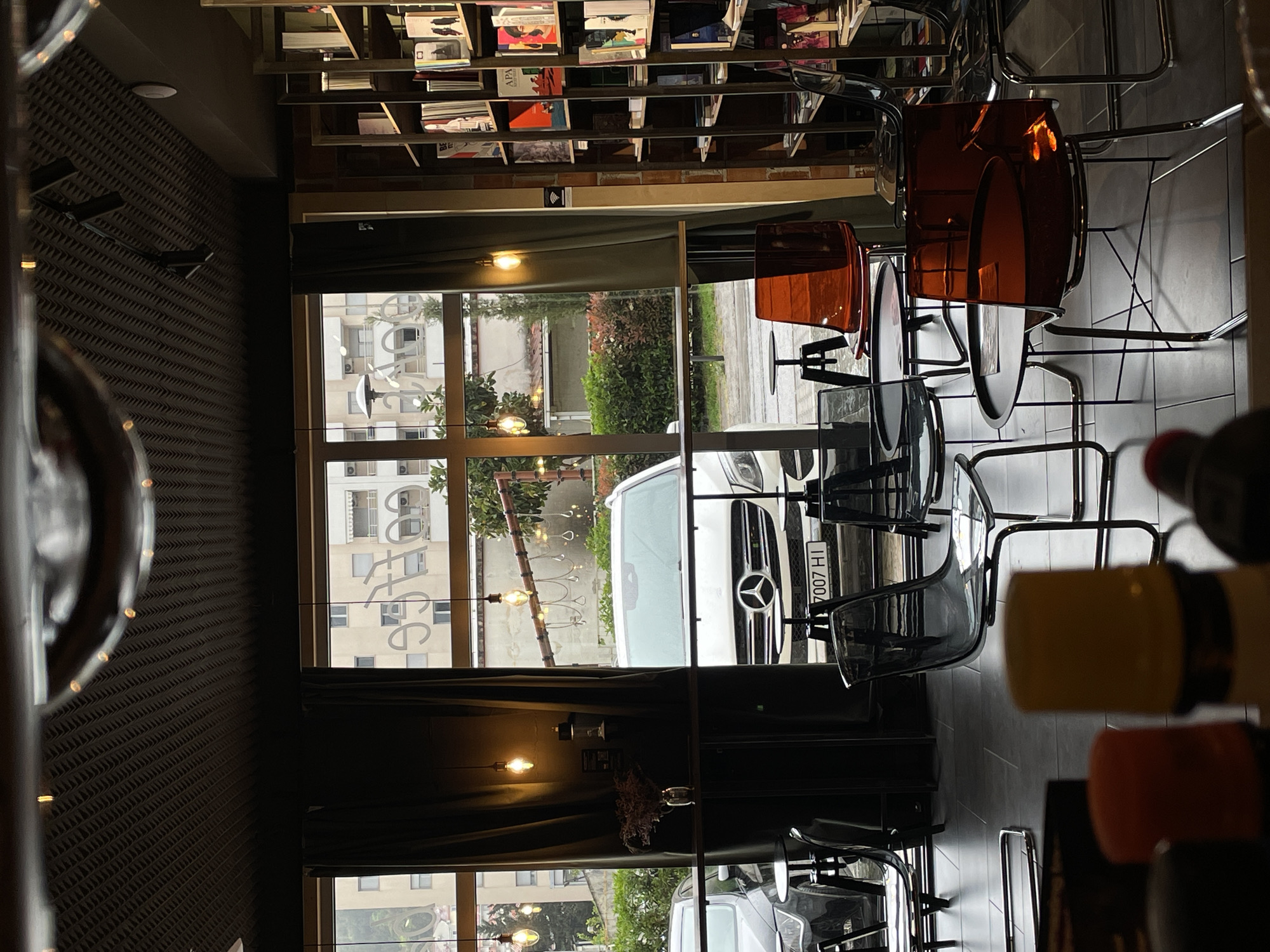 The image shows an interior view of a cafe or restaurant in Budva, Montenegro. A bookshelf filled with books runs along the top of the frame. Below, a window offers a view of the street outside, where a white Mercedes-Benz is parked. Reflected in the window is the word "COFFEE" written backward, suggesting it is displayed on the interior of the glass. There are modern chairs with orange and grey translucent backs and metal frames arranged in the foreground. The interior design includes dark, textured wall treatments. Through the window, urban buildings and greenery can be seen. The environment suggests a contemporary, urban setting within Budva.