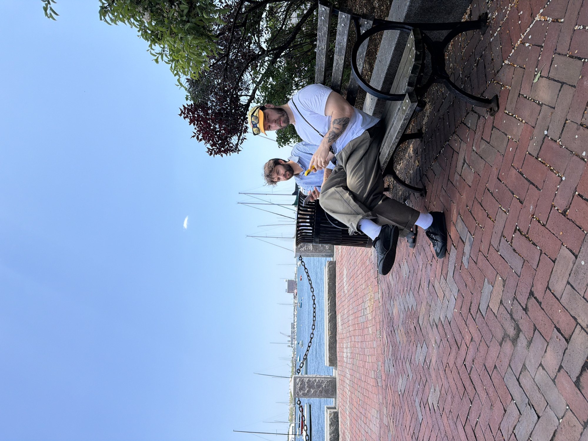 Two Caucasian males are seated on a park bench. The first male is seated to the right; he has facial hair and is wearing a white T-shirt, brown pants, and black shoes. He is also wearing sunglasses on top of his head. The second male is seated next to him; he has facial hair and is wearing a blue shirt, and khaki pants. He is holding a small yellow object in his hands. They are seated on a wooden bench next to a body of water, presumably a harbor. There are several sailboats in the background, and a chain fence runs along the edge of the waterfront. The ground is paved with red bricks. The location is likely Boston, United States of America.