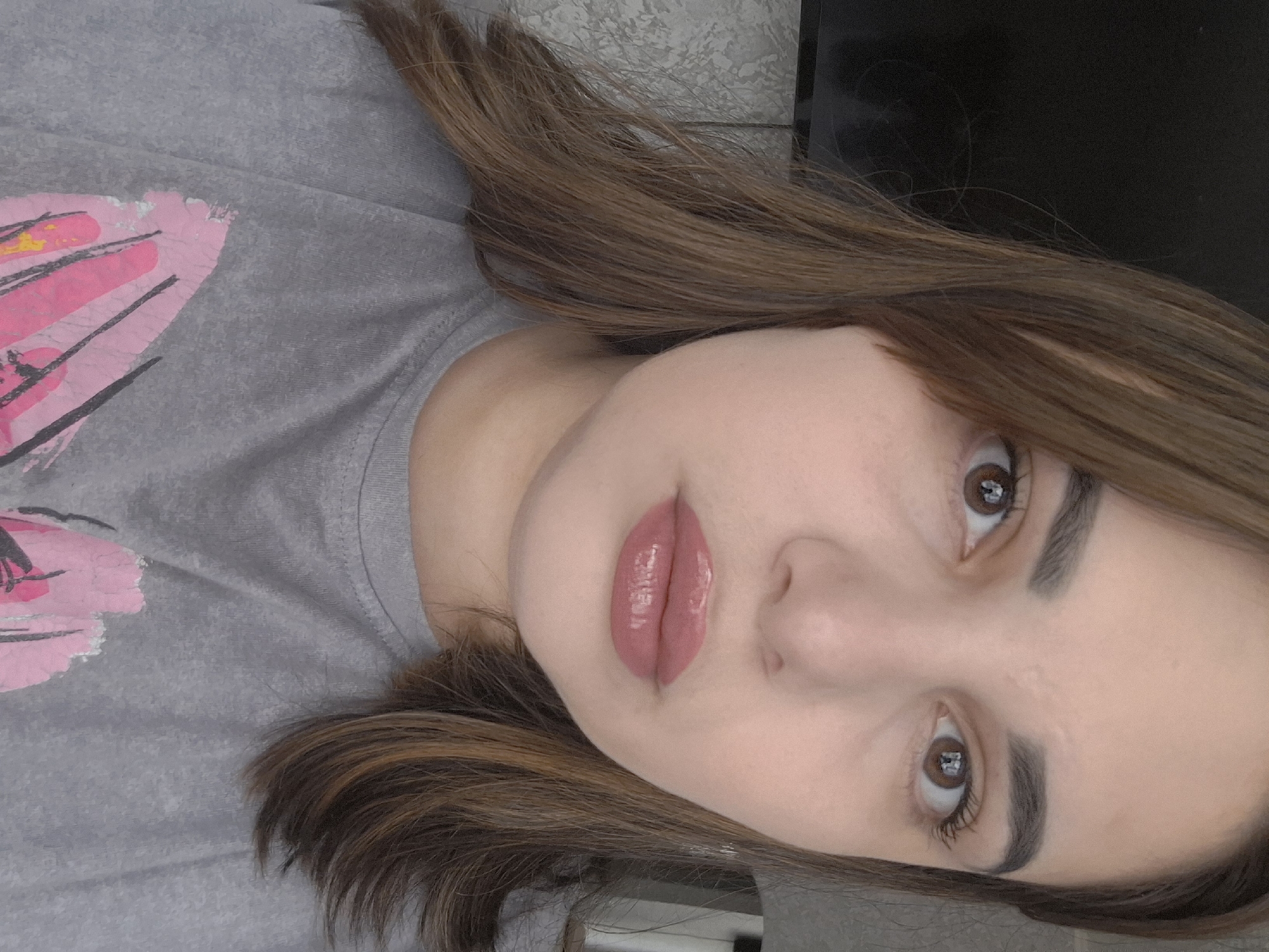 A young woman is pictured from a slightly elevated, angled perspective, likely a selfie. She has brown hair with highlights, brown eyes, and full lips coated in a glossy, pinkish lipstick. Her skin is fair. She is wearing a gray T-shirt with a pink graphic design on the left side of the frame.
The background is indistinct, with what appears to be a textured wall behind her and a dark, possibly electronic, object near the upper right corner.
There is no information in the image about the location being Chaikovsky, Russia.