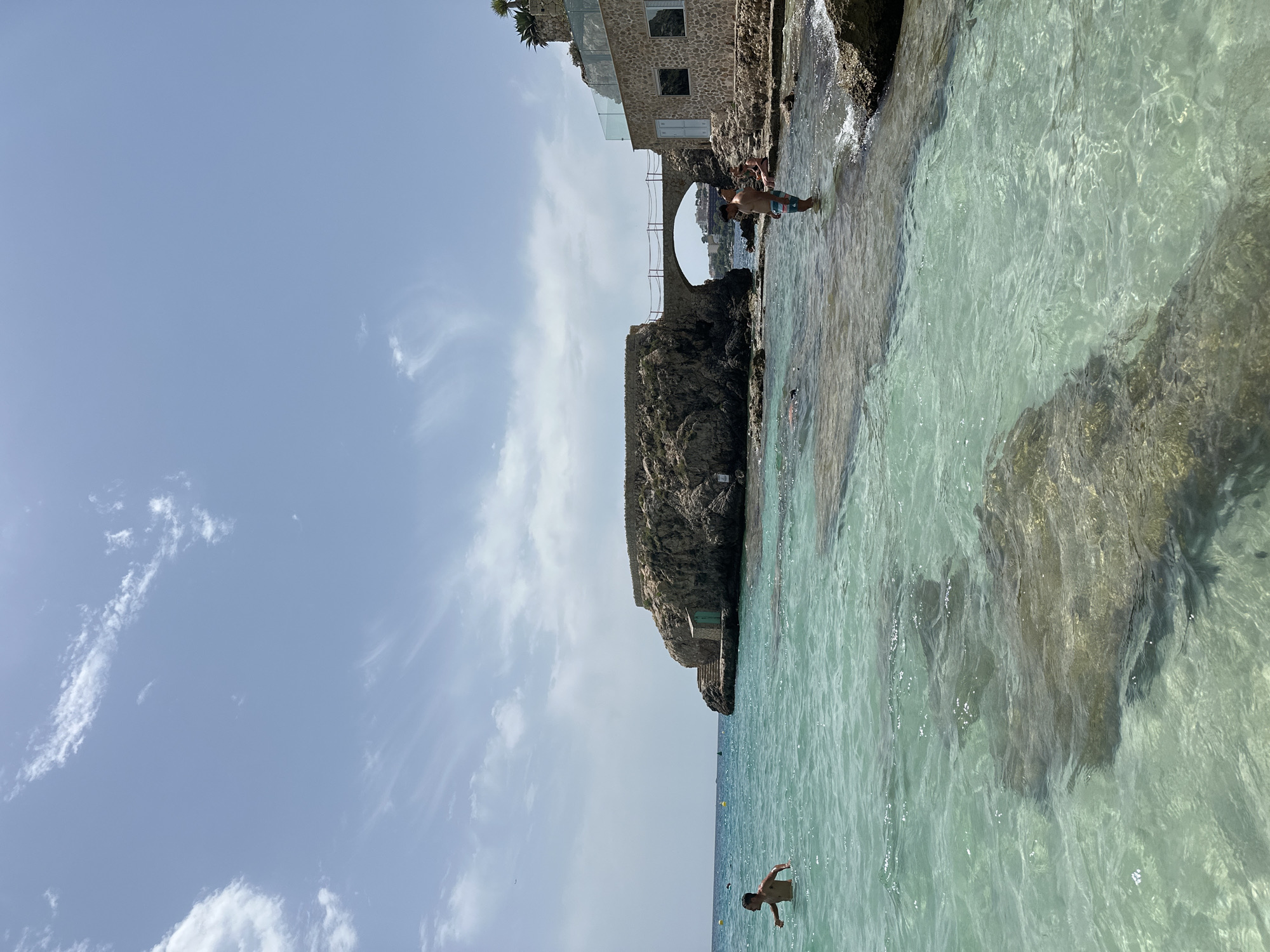In the city of Sant Agustí, Spain, on a clear day, a group of people enjoy the turquoise waters of a rocky cove. The scene unfolds with a clear blue sky dotted with wispy white clouds. In the foreground, the shallow, crystal-clear water reveals submerged rocks and sandy patches. Two young boys are visible in the water. One boy, wearing swim trunks, stands in the water, looking towards the rocky outcrop. Another boy is further out in the sea.
Dominating the middle ground is a rugged, rocky island with a small, green building constructed at its base, accessed by stone steps. A stone arch bridge connects this island to a coastal structure built with light-colored stone. The structure features arched windows and a balustrade overlooking the sea.
On the right side of the image, a man in swim trunks stands in the shallow water, facing left. Beside him, a woman sits on the rocks. The background shows the expanse of the sea meeting the horizon under the bright sky.