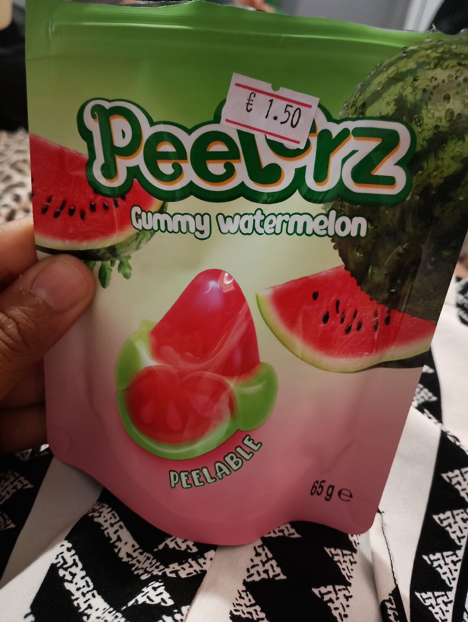 The image displays a close-up of a bag of "Peelz Gummy watermelon" candy. The bag is predominantly green and pink, featuring illustrations of watermelons and gummy candies shaped like watermelon slices. A price tag displaying "€ 1.50" is affixed to the upper right portion of the bag. A person's hand is visible on the left side of the image, holding the bag. The background consists of a black and white patterned fabric, likely clothing or bedding. The scene appears to be indoors. The image is a product shot of candy packaging, with no discernible elements suggesting a specific location in Zimnitsa, Bulgaria.