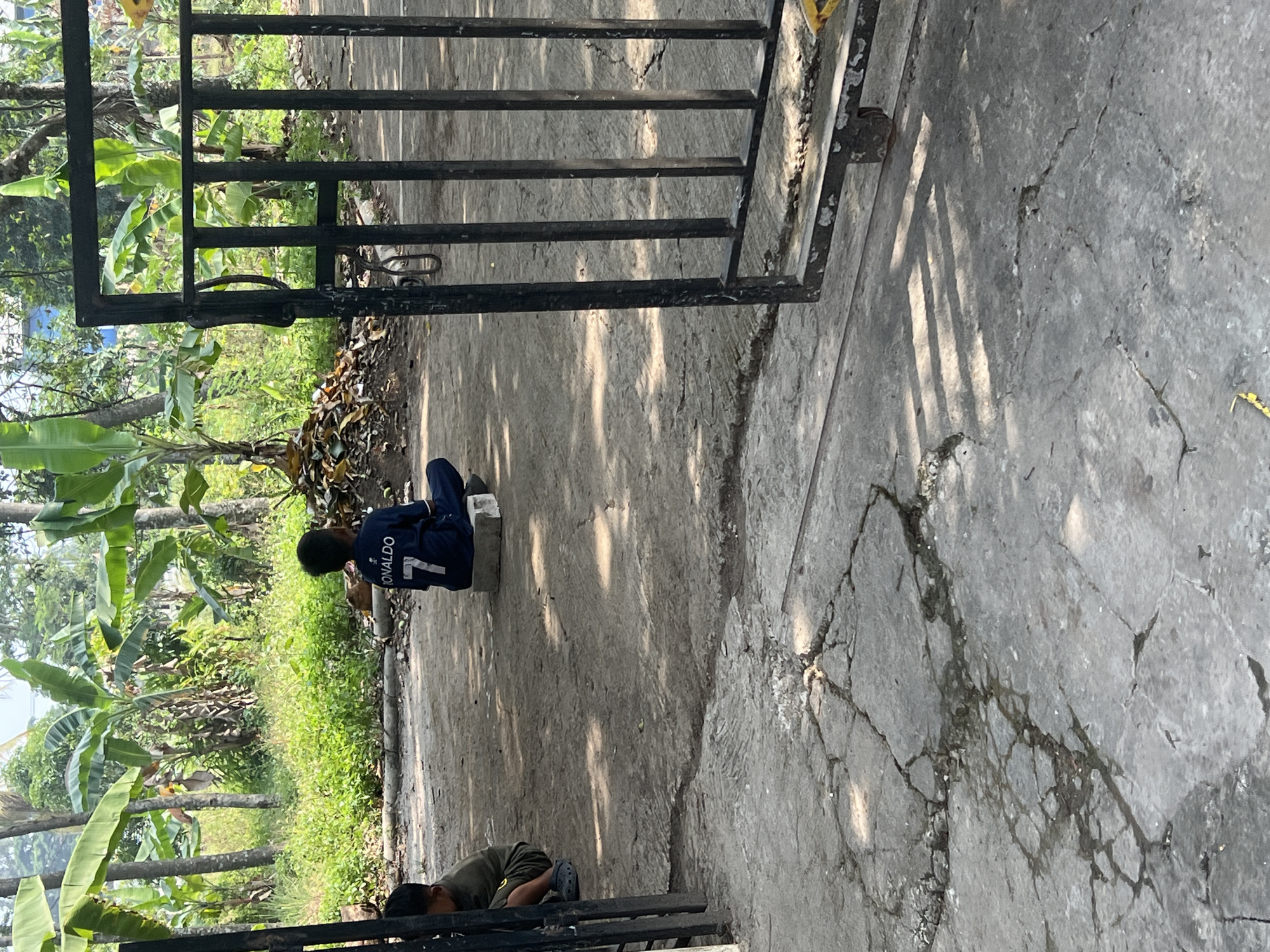 In the city of Klapanunggal, Indonesia, a child wearing a dark blue shirt with the name "RONALDO" and the number "7" is sitting on a concrete block in a yard. The child is facing away from the camera, looking towards a dense area of banana trees and other vegetation. Another child, partially visible, is sitting to the left, facing towards the right. The ground is made of cracked concrete. A black metal gate is visible on the right side of the frame, open. The scene appears to be outdoors in a residential or semi-rural area.