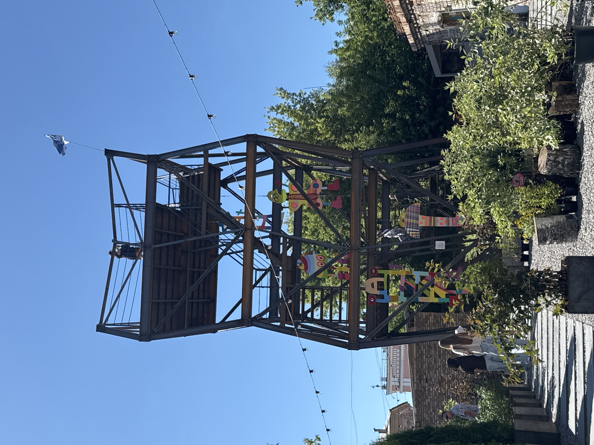 An outdoor scene features a tall, metal, lattice-like structure with multiple platforms and stairs. A flag flies from the top. Colorful, cartoonish figures are attached to the structure at various levels. In the foreground, people walk along a path lined with greenery in planters and decorative logs. The background shows brick buildings and trees under a clear blue sky. The location is identified as a park in the city of Yelets, Russia.