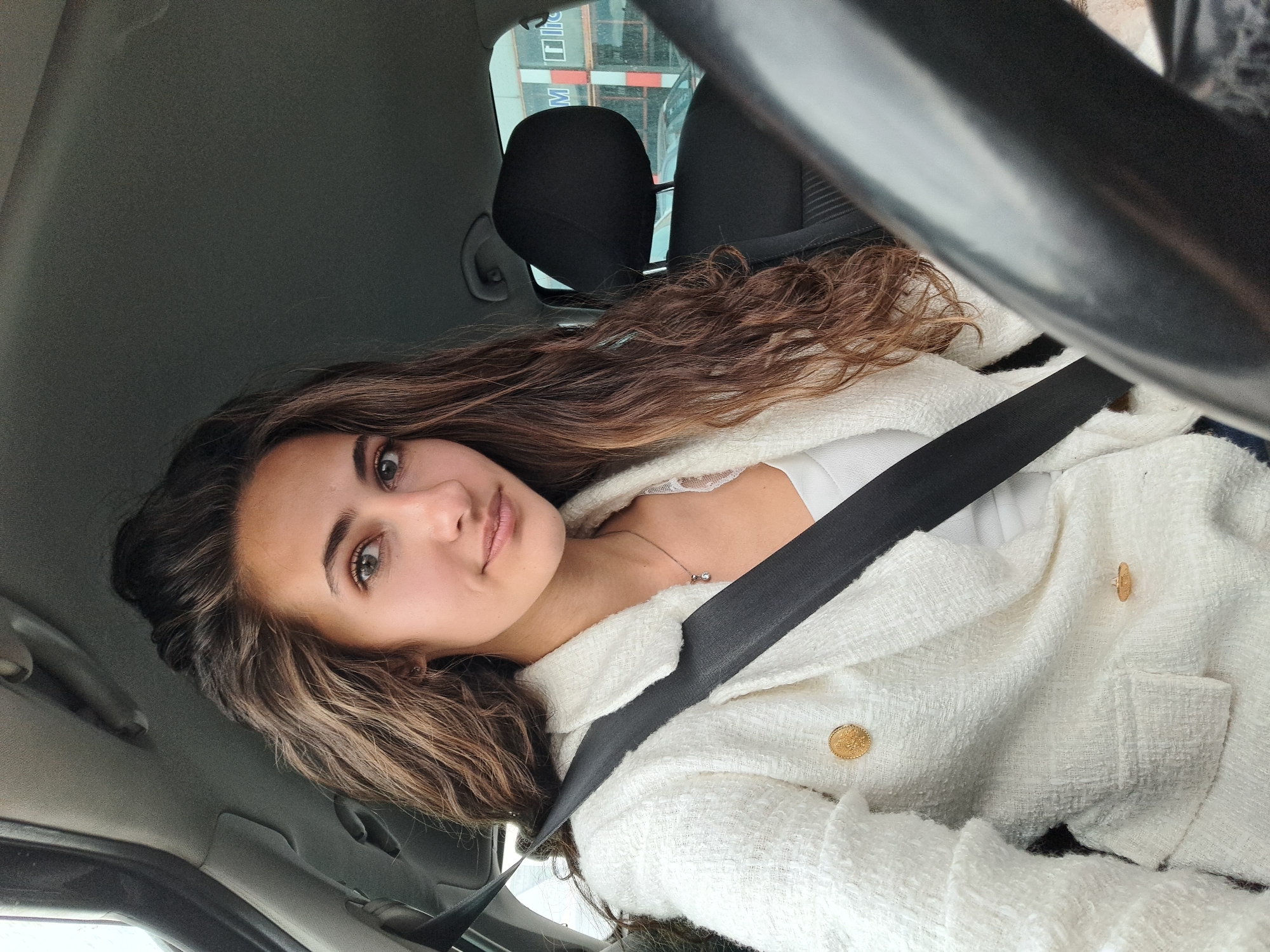 A young woman with long, curly brown and blonde hair is sitting in the driver's seat of a car, wearing a white textured jacket with gold buttons and a seatbelt. She is looking directly at the camera. The car interior is visible, including the steering wheel, headrest, and dashboard. The background shows a building with a glass facade, and elements of what seems like a construction site are visible outside the car window. There is no clear indication in the picture that directly places it in Sofia, Bulgaria.
