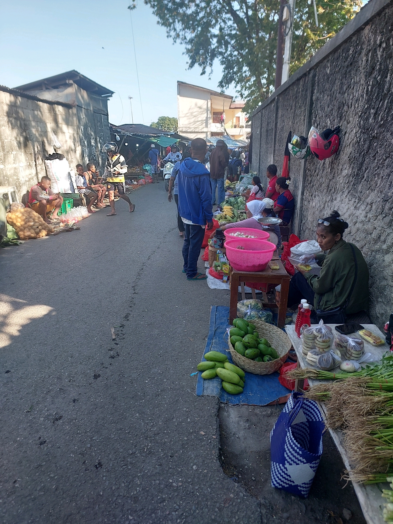 The image depicts a street market scene in Kupang, Indonesia, likely a local roadside market. The setting is an asphalt road with a rough, textured concrete wall on the right side. Various vendors are present, selling produce and other goods from makeshift stalls.
In the foreground on the right, a woman with glasses and her hair tied back is seated, looking towards the left. She is wearing a dark green jacket over a white shirt and is holding what appears to be food or a small item. Next to her, on a table covered with a cloth, are various packaged goods, including what look like cakes or snacks in plastic packaging, and some bottles. A basket filled with green mangoes is on the ground in front of the table, along with some loose green mangoes. A blue and white checkered bag is also on the ground. Further to the right, bundles of green vegetables with roots are displayed.
Along the wall, two red and green helmets are hanging. Other vendors are visible further down the street, some sitting and others standing. There are several people walking or standing around the market stalls. To the left of the road, a building with a corrugated metal roof and rough walls is visible, with a pile of sacks next to it. Several young males are sitting in front of this building, one of whom is smoking.
The overall atmosphere suggests a busy, informal marketplace under a clear sky, with sunlight casting shadows on the ground. The presence of local architecture, the style of the market stalls, and the appearance of the people are indicative of a scene in Indonesia.