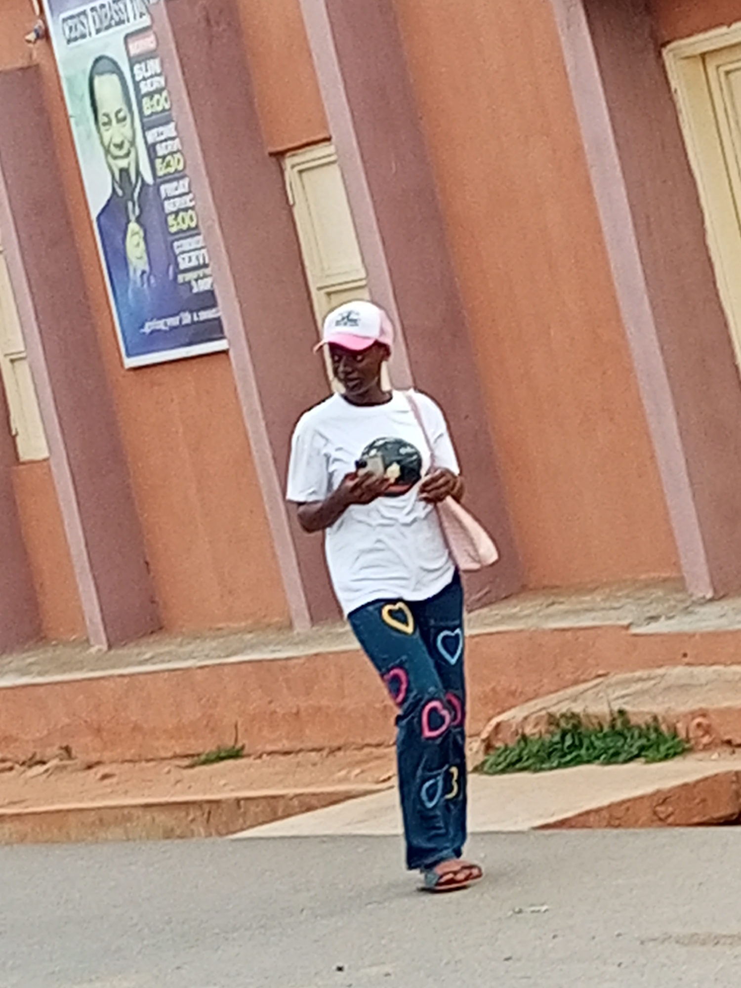 A young Black woman wearing a white t-shirt and blue jeans adorned with colorful heart patches, a pink baseball cap, and red sandals is standing on a paved street in front of a reddish-brown building. She is holding a smartphone in her right hand and a light pink handbag over her left shoulder. The building has a poster featuring a caricature of a man with a goatee, along with a schedule of events. The image appears to be taken during the day, with soft lighting. The overall setting suggests a street scene in Tempure, Nigeria, likely a commercial or public area given the building's facade and poster.