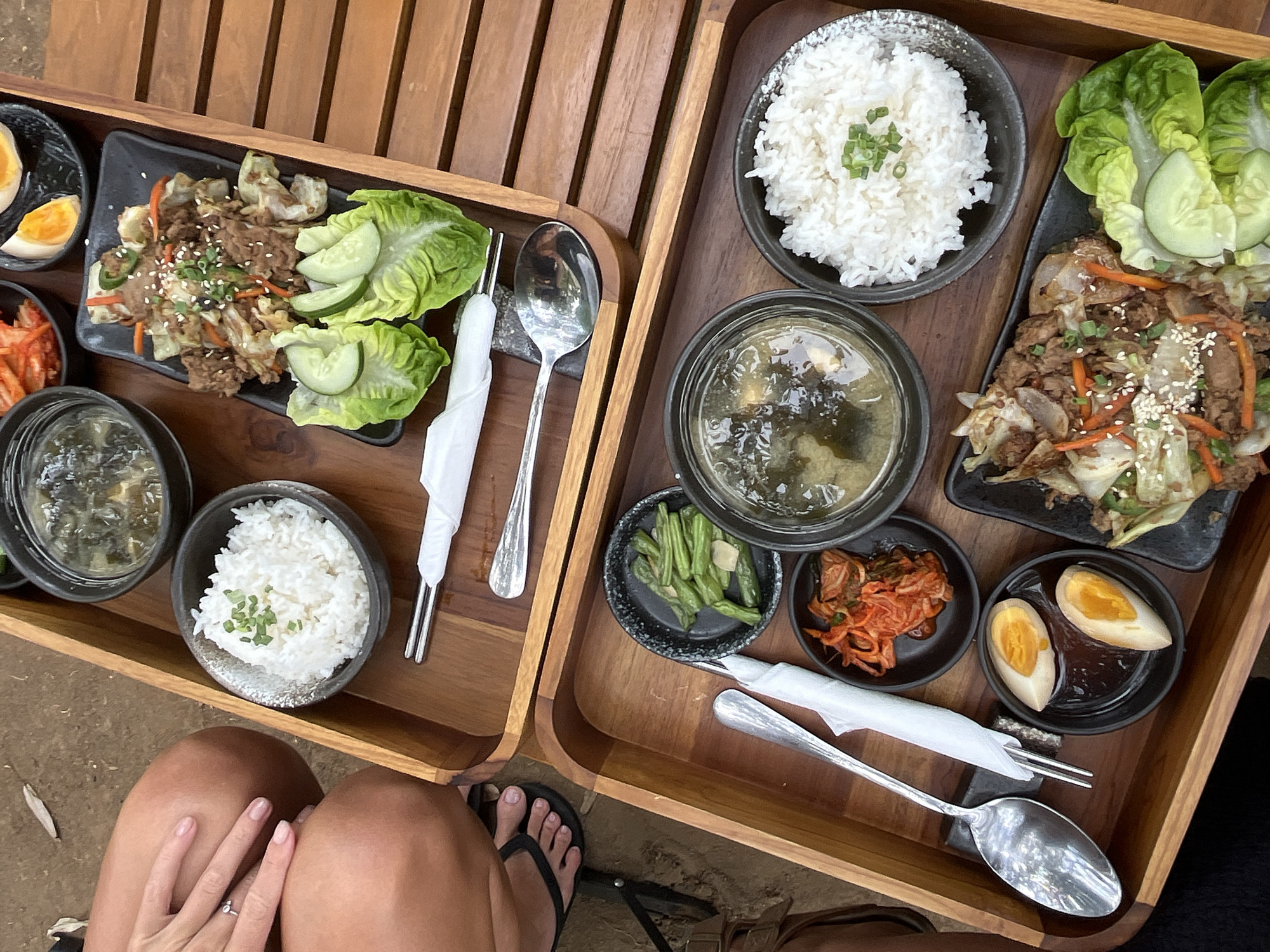 The image depicts two wooden trays filled with food, presumably in Kuta Selatan, Indonesia, given the context. The upper tray contains a bowl of white rice, a bowl of soup, a dish of stir-fried meat and vegetables garnished with sesame seeds and scallions, a small bowl of kimchi, and several slices of cucumber on lettuce leaves. Cutlery, wrapped in white paper, is also visible. In the lower tray, a similar arrangement includes a bowl of rice, a bowl of soup, green beans, kimchi, two halves of a hard-boiled egg, and a dish of stir-fried meat and vegetables with cucumber and lettuce. Cutlery is also present in this tray. A person's legs and feet, wearing flip-flops, are visible on the left side of the frame, suggesting they are seated and reaching for or interacting with the food. The background appears to be an outdoor setting with a sandy or earthy ground surface and wooden planks, consistent with a restaurant or dining area in a tropical location.