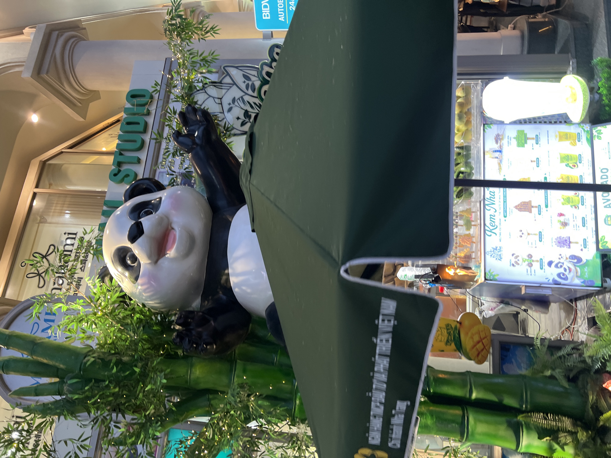 The image captures a lively street scene in Nha Trang, Vietnam, centered around an ice cream stall. Dominating the foreground is a large, whimsical panda statue waving cheerfully, positioned in front of a dark green umbrella canopy. Behind the panda, a prominent sign reads "NAIROOM STUDIO" in green lettering against a white background, suggesting a creative or retail space. The stall itself is brightly lit, displaying a variety of ice cream options with appealing images of popsicles and cups. A sign above the stall reads "Kem Nha" which translates to "Nha Trang Ice Cream," further pinpointing the location. The stall is adorned with bamboo-like decorations and features a large, stylized mango as a decorative element. The overall atmosphere appears festive and inviting, likely part of a commercial area or promenade in the city.