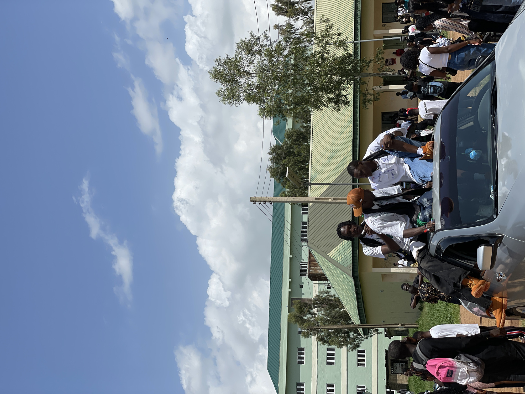 The image depicts a group of young people gathered outdoors, with a building and a car prominently featured. The scene appears to be set in a sunny, daytime environment, indicated by the bright sky with scattered clouds. The building has a light green exterior and a darker green roof, with multiple windows visible. The car is a light silver, and parts of its interior are visible through the windows.
The people are mostly dressed in casual attire, with a mix of t-shirts, jeans, and other everyday clothing. Several individuals are seated on the car, while others are standing around it. Some are looking towards the camera, while others appear to be engaged in conversation or observing something off-camera. One person in the foreground is wearing a white t-shirt with some writing on it and blue jeans, with a brown shoe on their foot. Another individual is wearing an orange cap.
The overall atmosphere seems to be relaxed and social, suggesting a casual gathering or event. There are no clear indicators of a specific organized event, but the presence of multiple people suggests a communal activity. The location appears to be an open area with some greenery, possibly a campus or public space.
While there is some text visible on the white t-shirt in the foreground, it is not clearly legible in the provided image. The general context suggests a scene in Wumat, Nigeria, under a bright, partly cloudy sky.