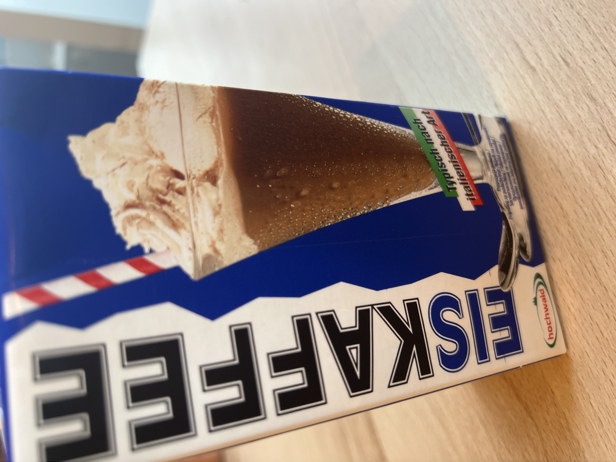 The image shows a close-up of a carton of "EISKAFFE" (iced coffee) manufactured by "hochwald". The carton features a blue and white design with large black letters spelling out "EISKAFFE" vertically. A glass of iced coffee topped with whipped cream and a red and white striped straw is depicted on the right side of the carton. Below the drink, text in green and red reads "Typisch nach italienischer Art" (typically in the Italian style). A small logo for "hochwald" is visible at the bottom left. The carton is placed on a wooden surface, likely a table. The background is blurred, suggesting an indoor setting. There are no people or specific landmarks visible in the image, and therefore no information can be deduced about its location in Vilnius, Lithuania.