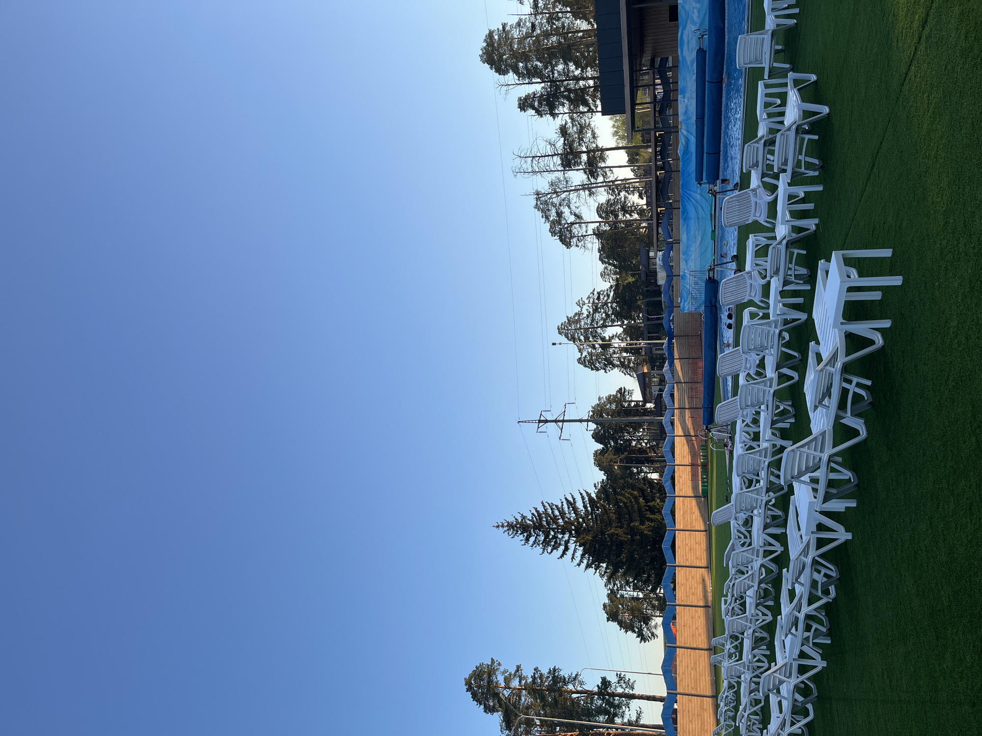 The image shows a daytime scene at what appears to be an outdoor pool area in Votkinsk, Russia. The sky is clear and blue. To the right are rows of white plastic lounge chairs arranged on green artificial turf. Behind the chairs is a pool area with a blue wave-patterned wall. Trees are visible beyond the pool area, along with utility poles and wires. The composition suggests the photo was taken in the late afternoon or early evening based on the lighting. There is no visible activity in the pool or on the chairs. The overall impression is a quiet or closed pool facility.