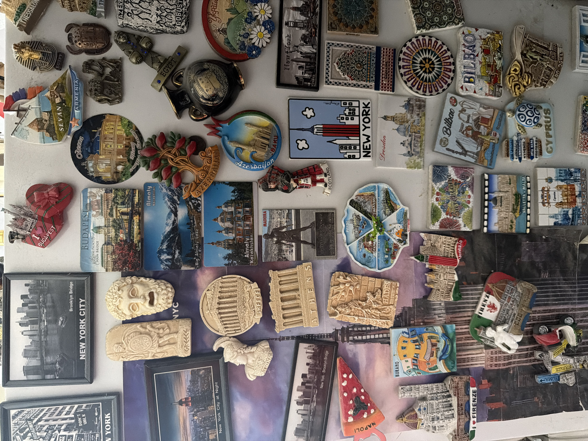 This is a vertically oriented, close-up photograph of a densely packed collection of souvenir refrigerator magnets, likely displayed on a metal surface such as a refrigerator door in Tbilisi, Georgia. The magnets, varying greatly in size, shape, and material, are arranged haphazardly, with many overlapping.
The collection primarily consists of travel-themed magnets representing numerous cities and countries worldwide. Subjects include iconic landmarks, city skylines, cultural symbols, historical figures, and artistic interpretations. There are flat, photographic, illustrative, and three-dimensional sculpted magnets made from materials like ceramic, resin, and metal.
Notable details include a wide geographical spread. Visible text and imagery identify locations such as:
* **North America:** NEW YORK CITY (with multiple magnets featuring the Brooklyn Bridge, Empire State Building, and skyline, as well as a "BROADWAY" sign).
* **Europe:** BUDAPEST ("Royal Palace & Chain Bridge"), Odessa (with "meamy opera baleta"), Dresden, SCOTLAND (a kilted figure), BILBAO (multiple), VALLETTA-MALTA, BURANO, NAPOLI (a pizza slice), FIRENZE (Florence, multiple), PISA, ROMA, VENEZIA (Venice, multiple), THESSALONIKI, Barcelona, Bologna. A magnet with a Ukrainian flag and "Україн" is also visible.
* **Asia/Middle East:** ARMENIA ("Sevan"), Almaty Kazakhstan ("Big Almaty Lake"), Azerbaijan ("Baku", pomegranate-shaped), CYPRUS (listing cities like Larnaca, Ammochostos, Kyrenia, Ayia Napa, Protaras, Limassol), EGYPT (a pharaoh bust and a cross-shaped magnet).
* **Other:** MADEIRA (featuring Cristiano Ronaldo), and Greece (with "ATHENAS," "GREECE," and a "HADES" magnet).
No specific activity is taking place; the image captures a static display of collected items. The lighting appears even, suggesting an indoor setting without strong natural light cues. The overall impression is one of extensive travel and a personal collection of memories.