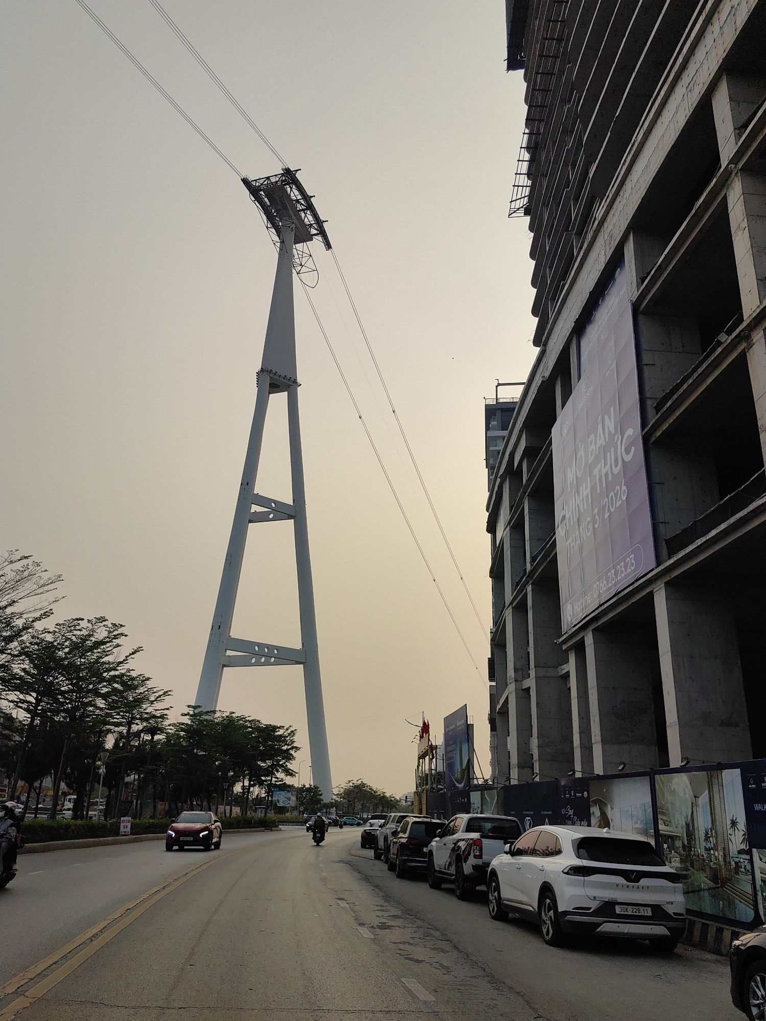 A street scene in Bai Chay Ward, Vietnam, features a large, white cable car pylon on the left side of the road, with cables extending diagonally across the frame towards the right. Cars and motorcycles occupy the road, while a line of parked vehicles sits along the curb. A partially constructed, high-rise building stands on the right side, with a large banner displayed on its façade. Trees line the left side of the road. The sky is overcast and a muted color.