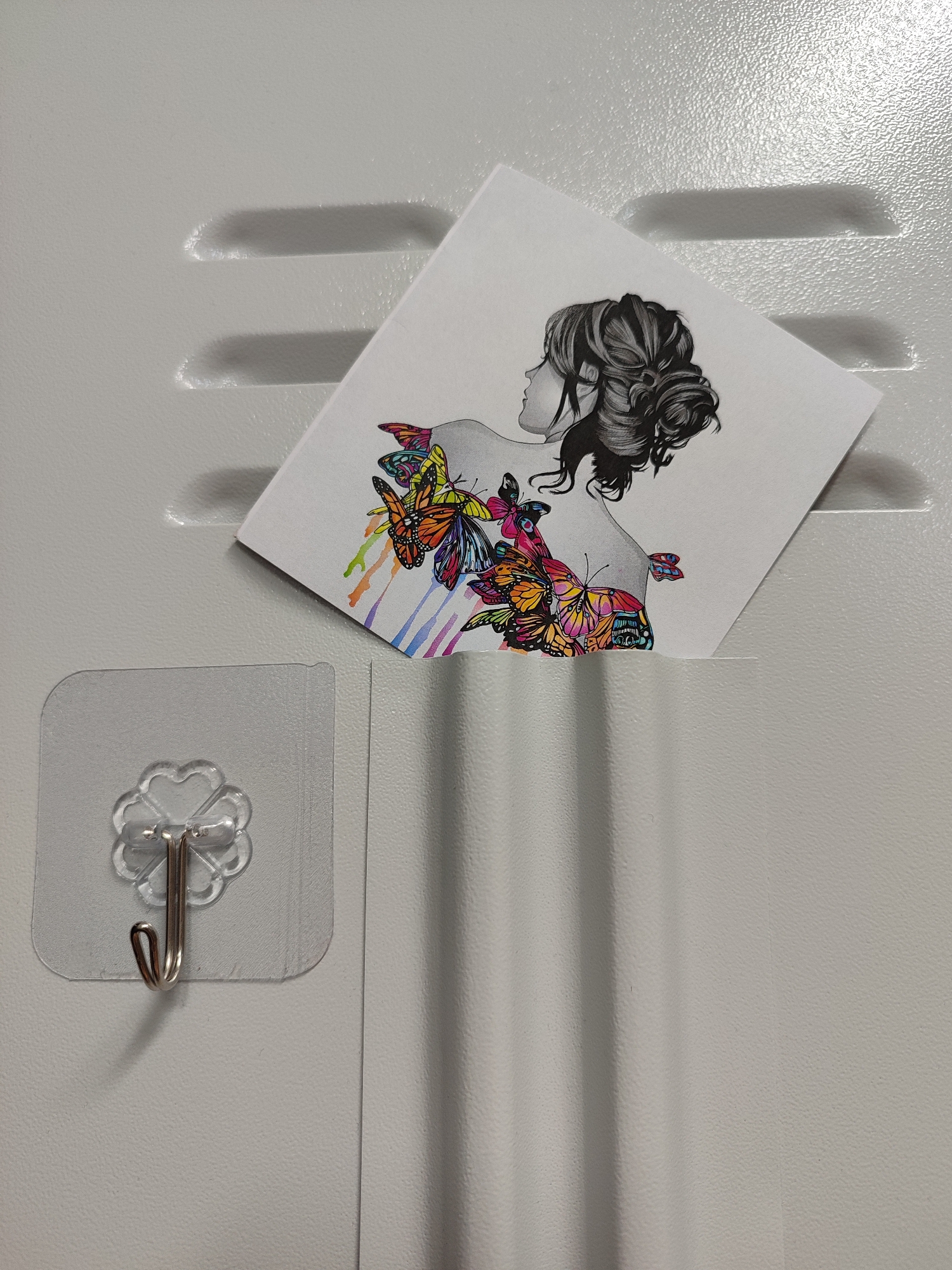 A black and white drawing of a woman's profile is shown, with colorful butterflies emerging from her back and cascading down her shoulder. The drawing is placed on a white, textured surface that has horizontal indentations. To the left of the drawing, a clear, flower-shaped adhesive hook with a silver metal hook is attached to the same textured surface. The overall scene suggests an indoor setting with a light-colored, possibly metallic or painted, surface.