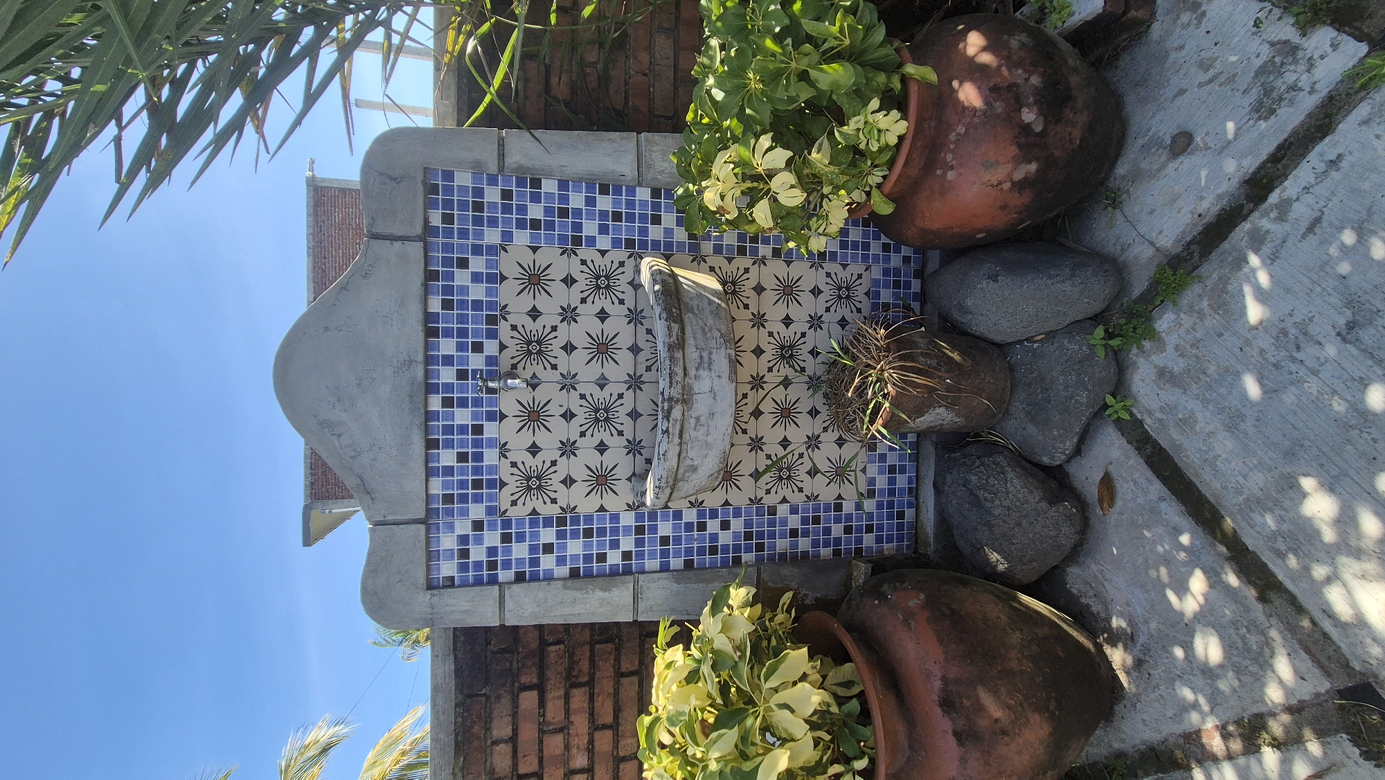 The image shows an outdoor fountain in Pekutatan, Indonesia. The fountain is decorated with blue and white mosaic tiles. A white, curved trough sits in the center of the tiled area. Two large, terracotta pots flank the fountain, each containing leafy plants. The fountain is positioned against a brick wall. The sky is visible in the background. The ground is paved with concrete and features natural stones. Some foliage is visible around the edge of the frame.
