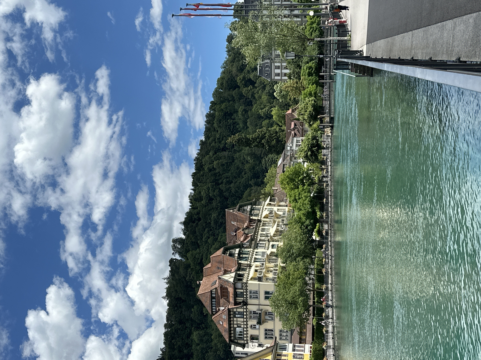 This is a daylight, eye-level, medium shot of the Aare River in Thun, Switzerland, from the promenade. The river occupies the majority of the lower half of the frame and displays a turquoise hue, with ripples and reflections of the sky and surrounding buildings. The promenade runs along the right side of the image, featuring a black metal railing, a paved walkway, and a few scattered people.
On the left side of the river, a row of buildings with traditional Swiss architecture lines the waterfront. The most prominent building is a large, pale yellow structure with a reddish-brown tiled roof, featuring numerous windows and balconies. Behind these buildings, a densely forested hill rises steeply. The sky above is a vibrant blue, dotted with fluffy white clouds.
Several people are visible walking or cycling along the promenade and the riverbank. The scene conveys a serene and picturesque atmosphere, typical of a summer day in a Swiss city.