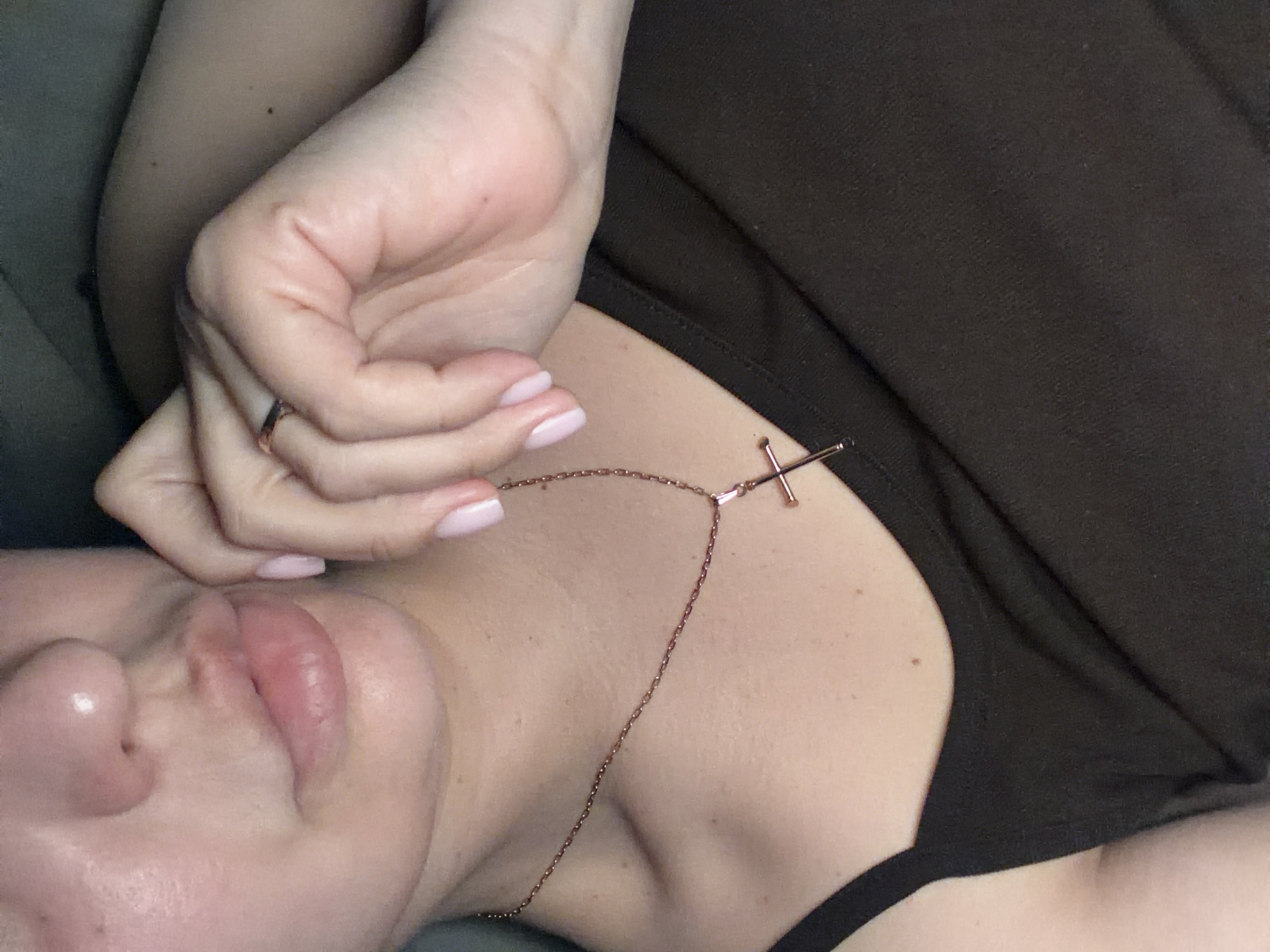 A close-up, angled photograph captures a person (likely female) reclining indoors, with a focus on their upper body, face, and hand. The subject has fair skin with a scattering of small moles or freckles visible on the chest and shoulder area. Their hand, adorned with a delicate gold or rose gold ring on the ring finger, features light pink, oval-shaped manicured nails. This hand rests near the person's jawline, partially obscuring the lower face, though full lips and a portion of the nose are visible. The person wears a slender, rose gold-colored chain necklace with a simple, bar-style cross pendant that lies on their collarbone and upper chest. A dark, possibly brown or black, garment with visible fabric texture covers the upper torso and shoulder. The background appears to be a muted, dark fabric, suggesting an upholstered surface like a couch or bed. The lighting is soft, creating an intimate and relaxed atmosphere. There is no visible text in the image. While the location context is given as Sochi, Russia, there are no visual cues within the image to confirm this specific setting.
